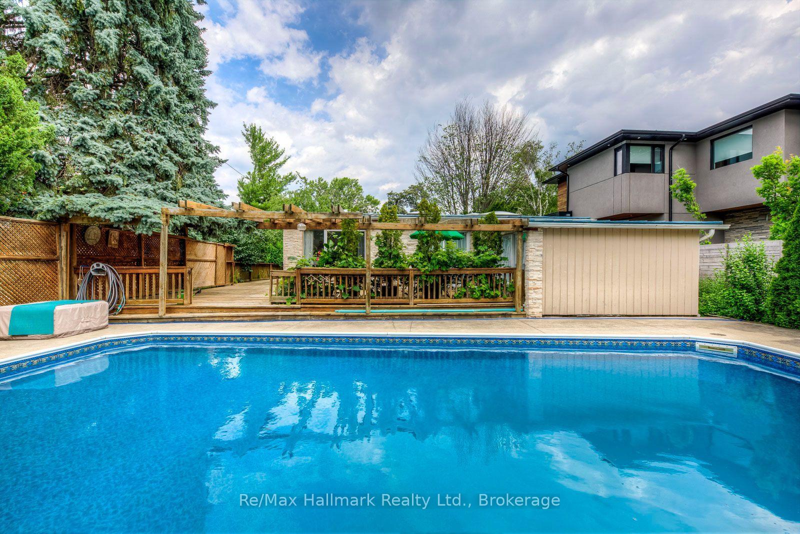 Backyard swimming pool beside a wood deck, pergola, and mature evergreen trees.