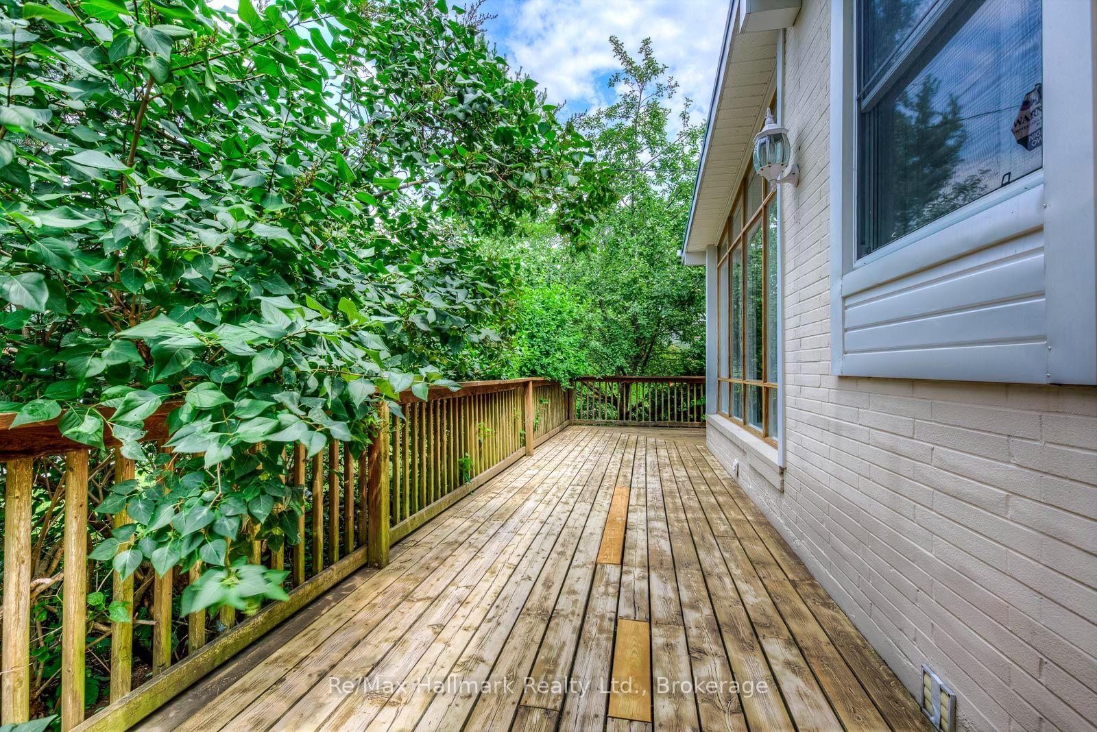 Wooden deck beside a white brick house, bordered by a railing and dense leafy greenery.