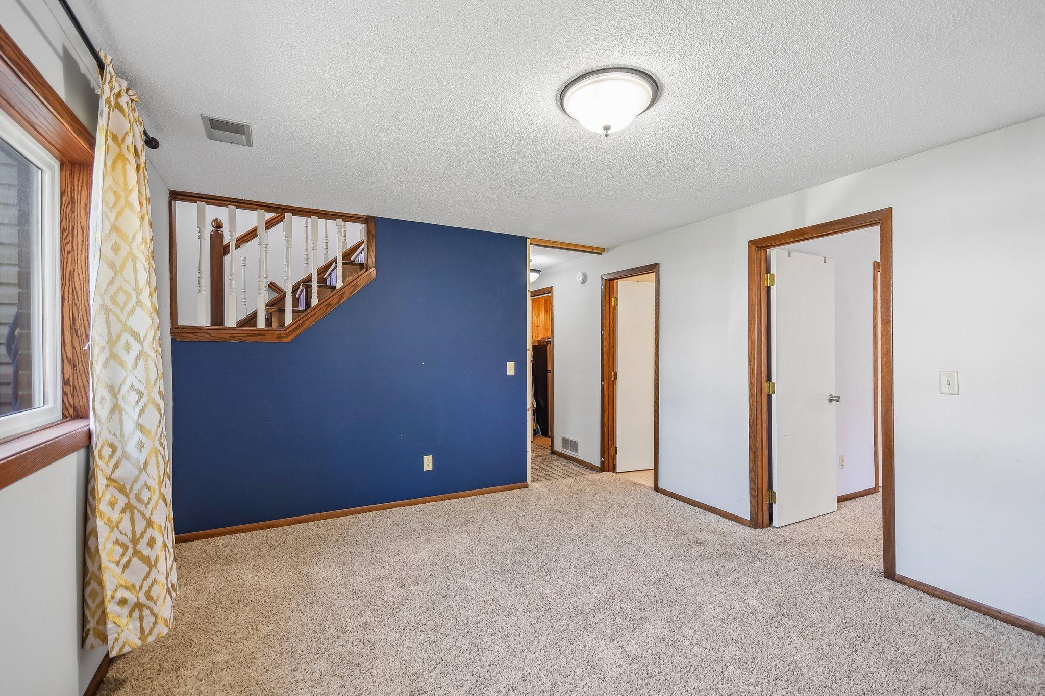 Empty carpeted room with a blue accent wall and wood-trimmed staircase railing.