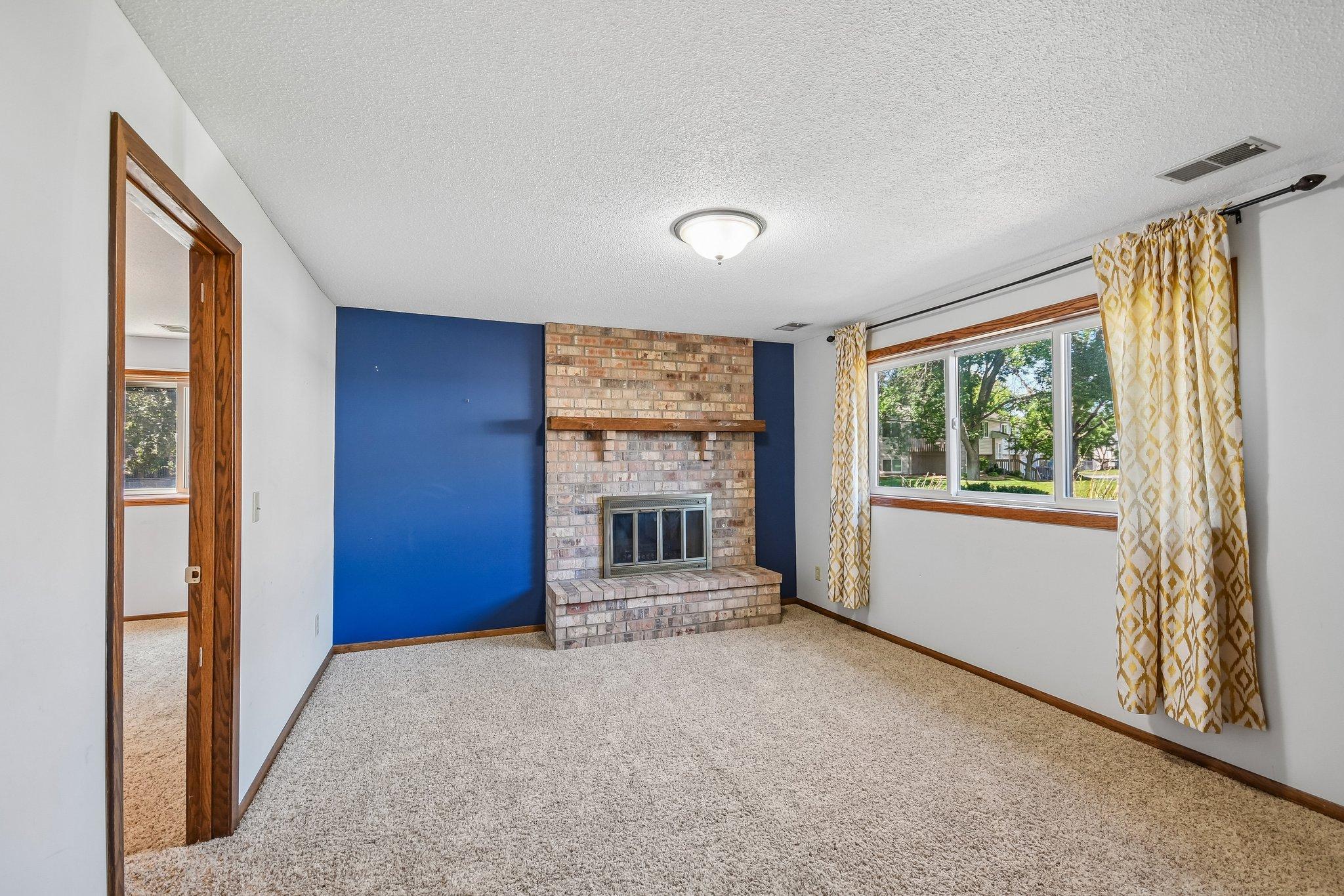 Empty living room with a brick fireplace, blue accent wall, and wide curtained window.