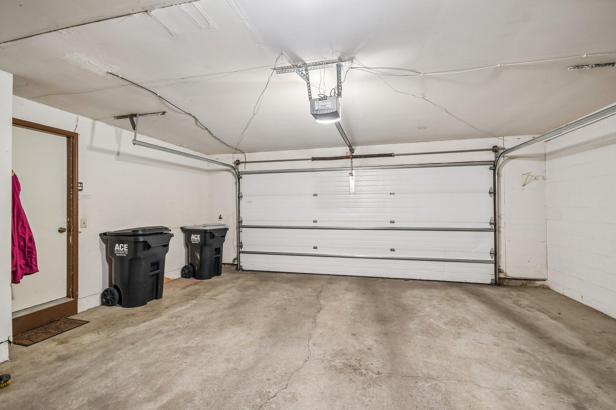 Garage with a closed sectional door, concrete floor, ceiling-mounted opener, and two black bins.