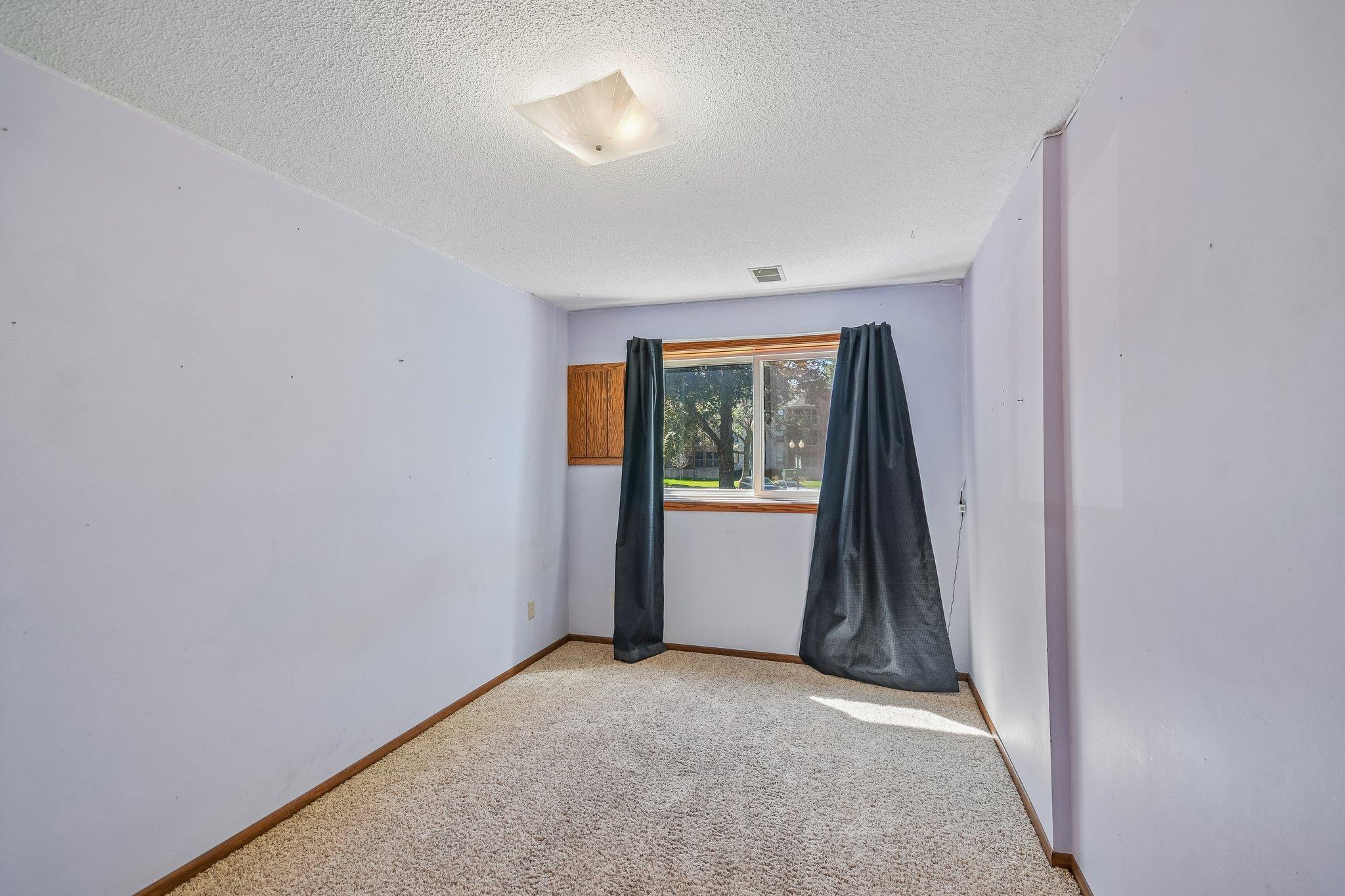 Empty carpeted room with lavender walls, dark window curtains, and sunlight on the floor.