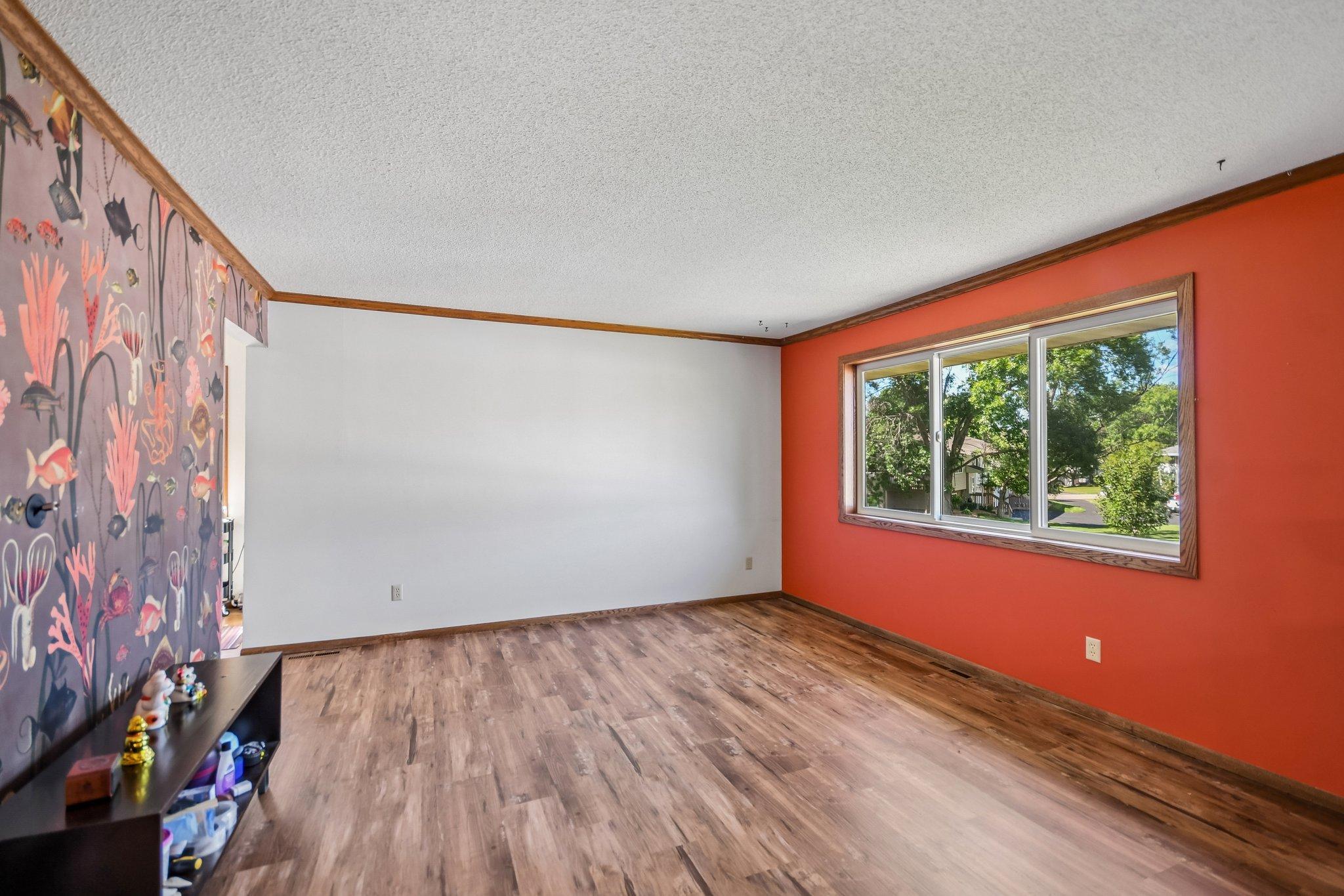 Mostly empty room with wood-look flooring, a rust-red wall, and underwater-themed wallpaper beside a large window.