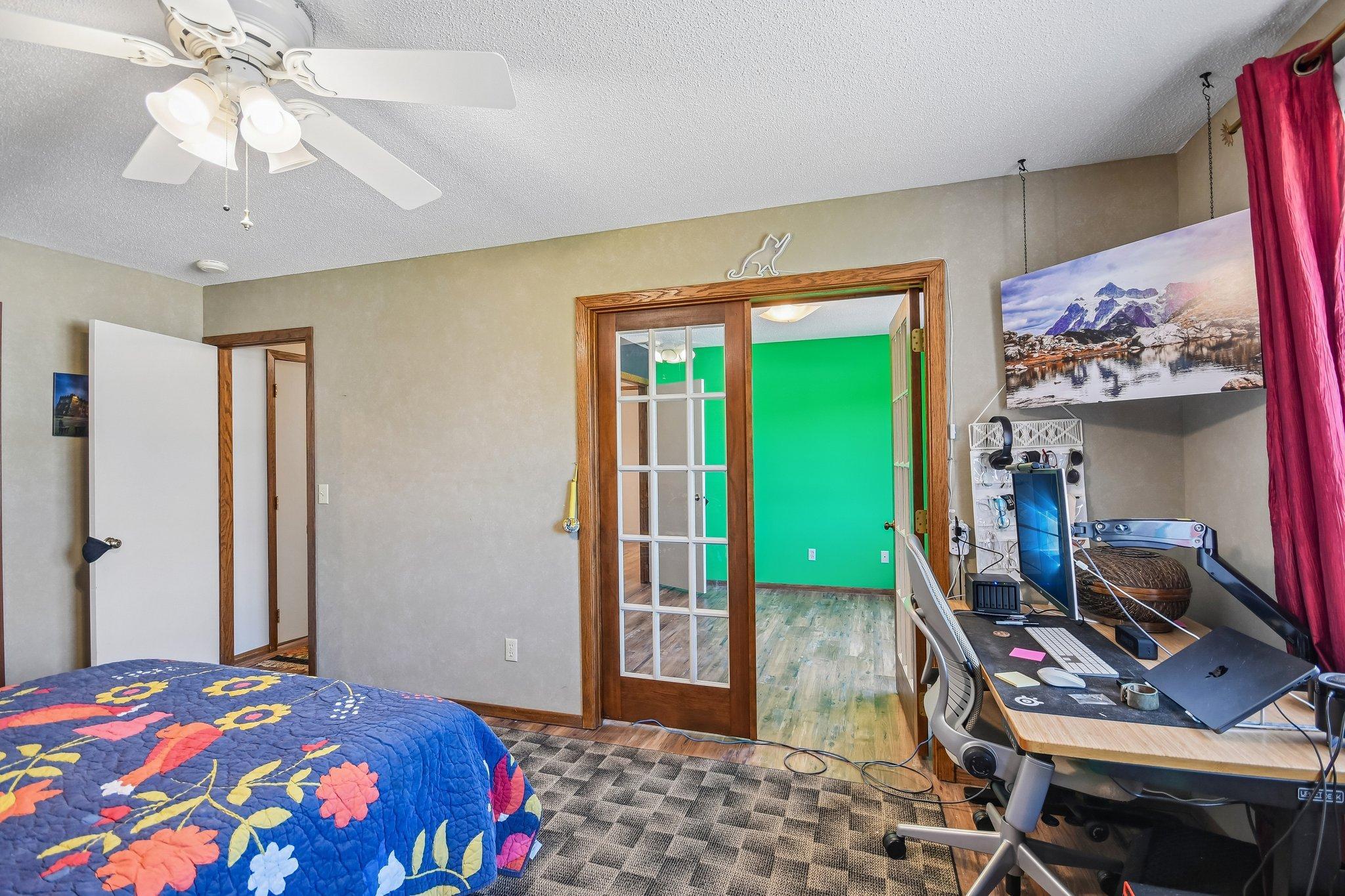 Bedroom with floral quilt and computer desk; French doors open to a bright green room.