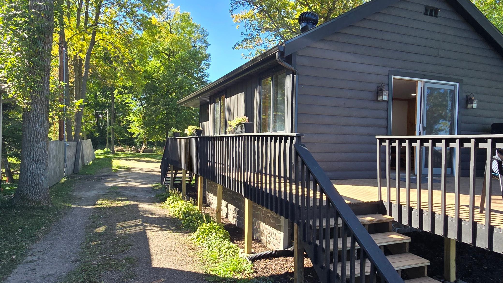 Dark-sided house with raised deck and stairs beside a wooded yard and gravel path.
