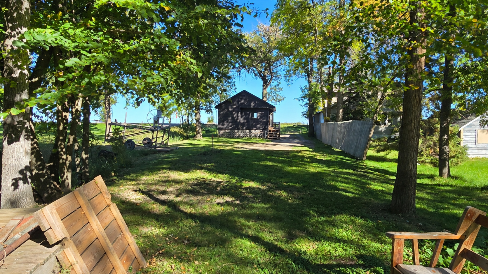 Tree-shaded yard with a small dark building, stone foundation, and wooden chairs.
