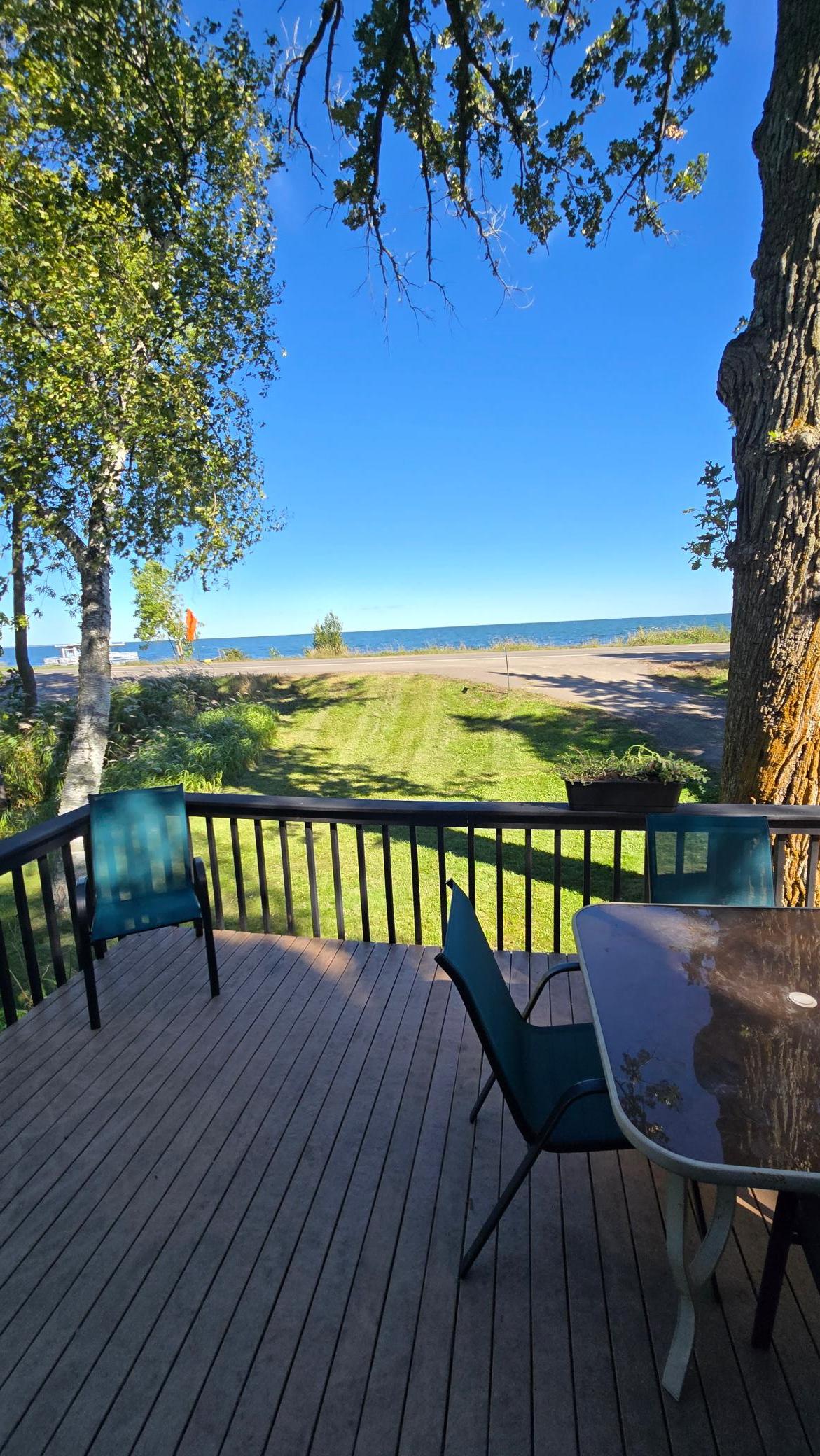 Deck with patio seating overlooking a grassy yard, roadside, and lake.