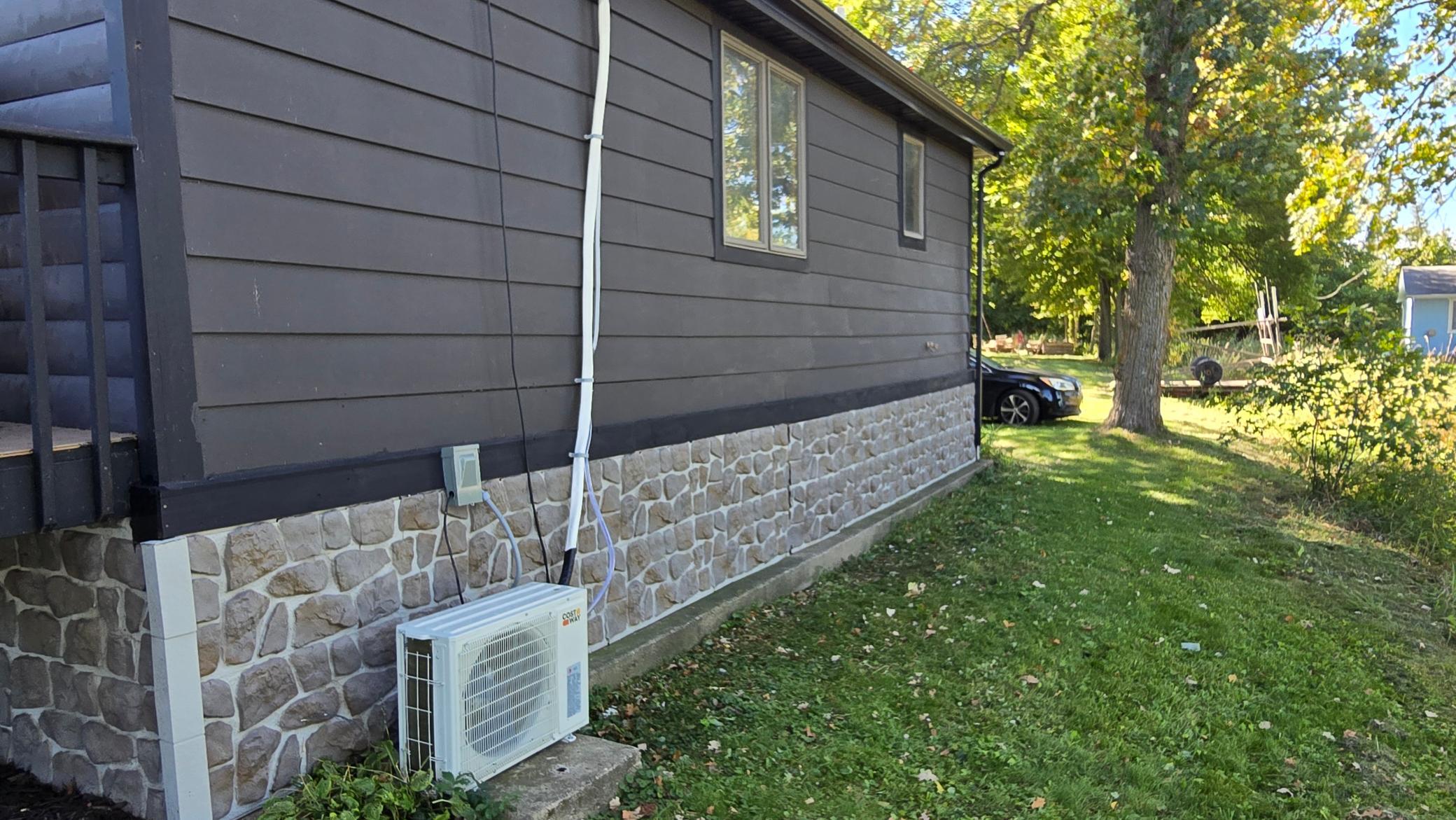 House side with stone foundation and outdoor heat pump beside a tree-lined lawn.