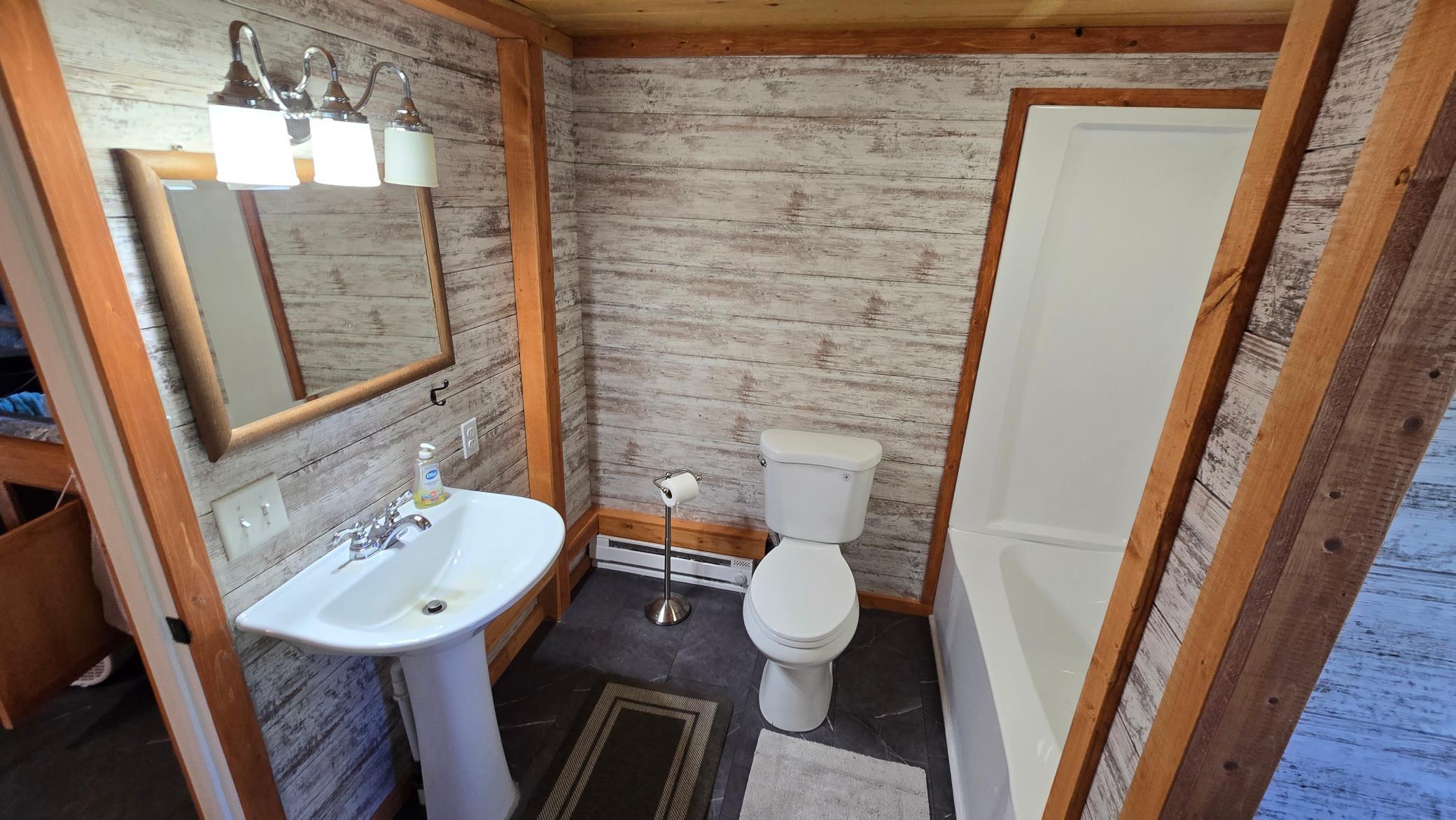 Bathroom with weathered plank walls, a pedestal sink, toilet, and bathtub-shower combination.