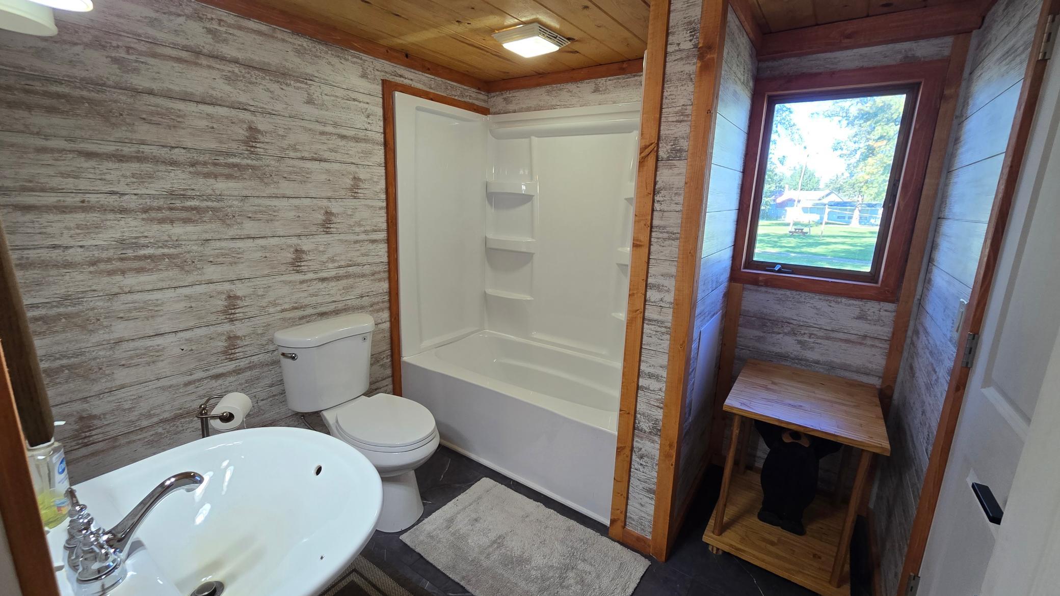 Bathroom with a white tub-shower and toilet, weathered plank walls, and a windowed wooden bench nook.