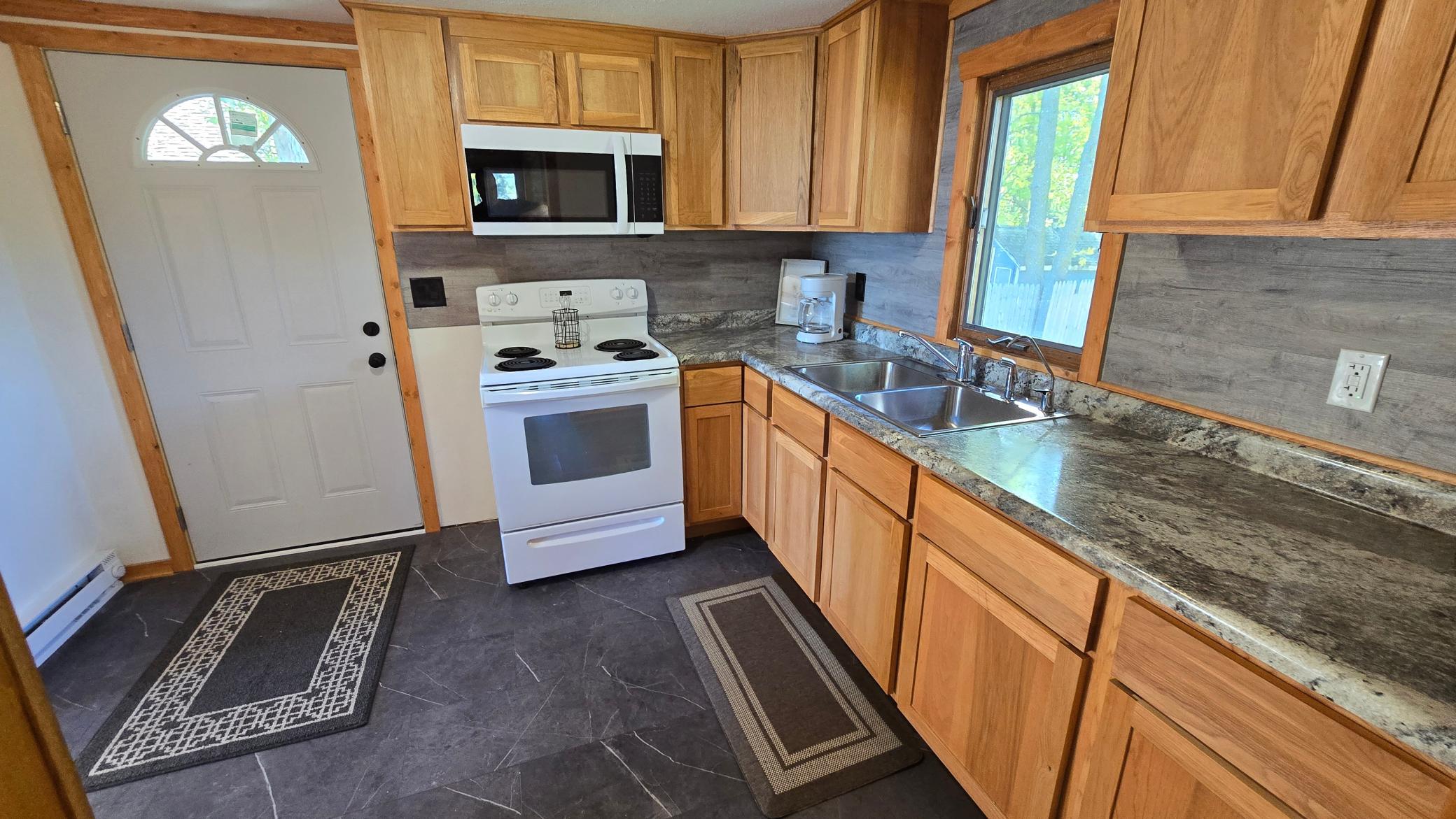 Kitchen with honey-toned wood cabinets, gray counters, white appliances, and a double sink beneath a window.