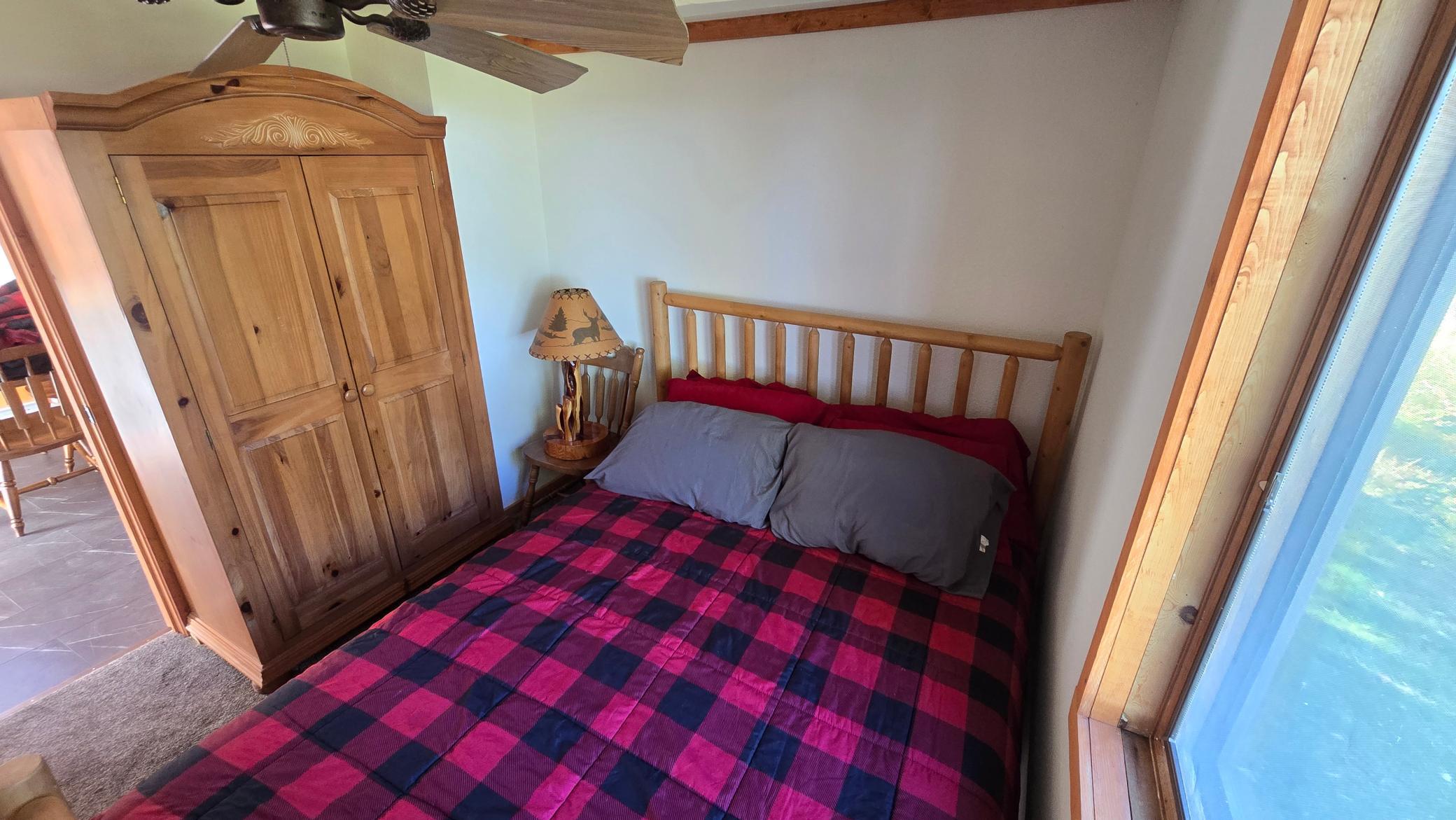 Bedroom with a red-and-navy plaid quilt, pine bed and wardrobe, and a large window.