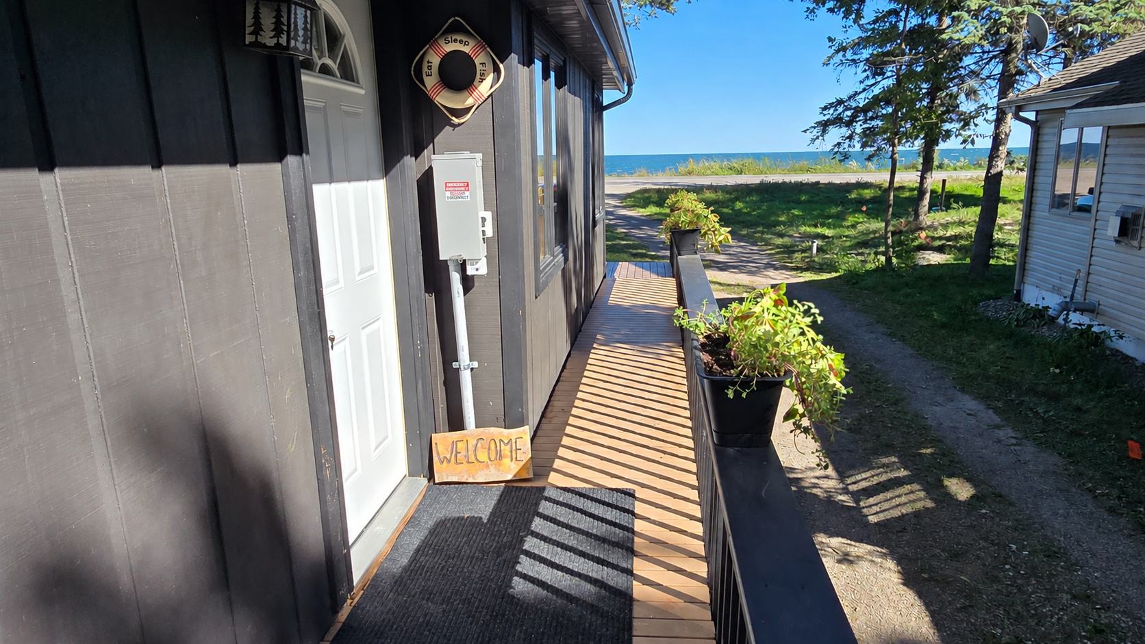 Lakeside property entrance with a wooden walkway, potted plants, and water visible beyond the yard.
