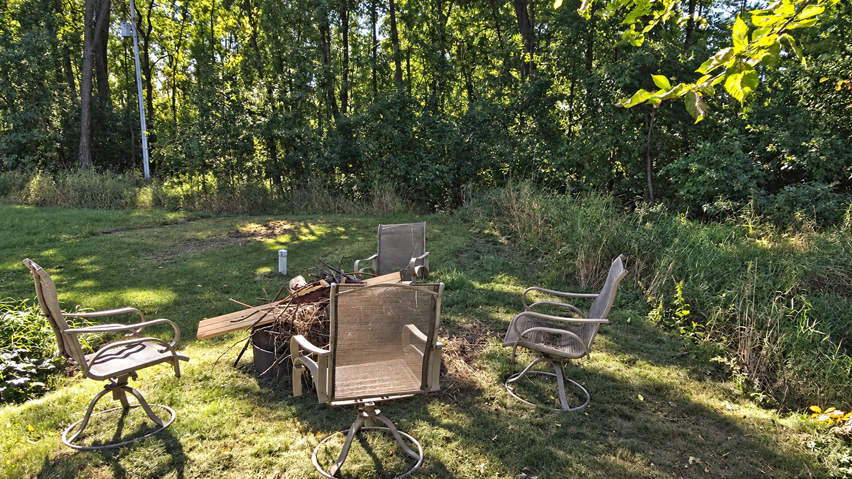 Wooded backyard seating area with patio chairs around a fire pit.