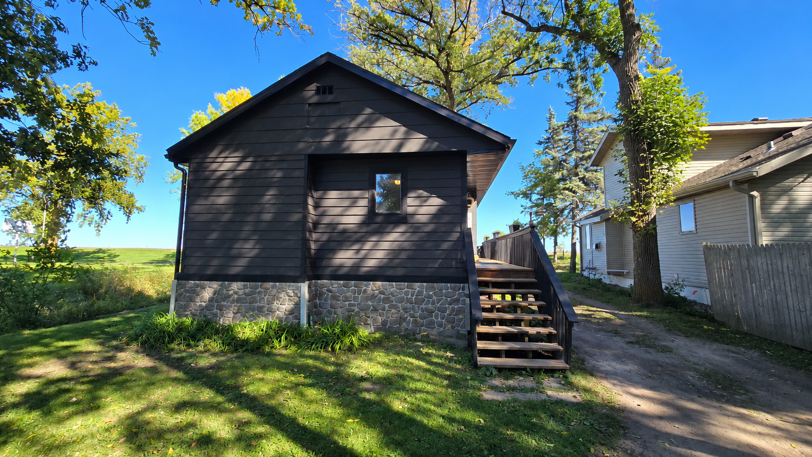 Dark-sided house with a stone foundation and wooden entry steps beneath mature trees.