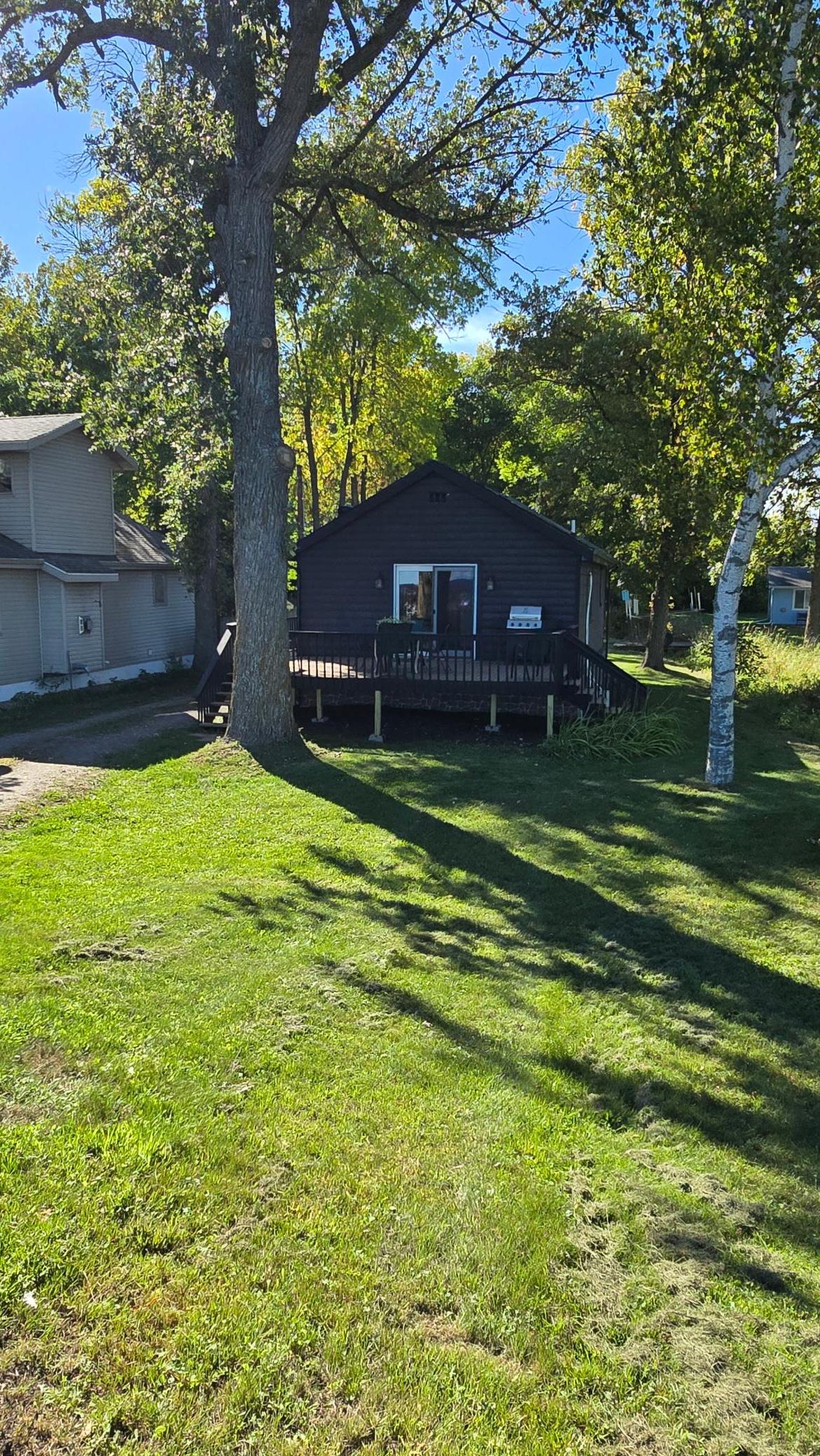 Dark-sided cabin with a raised deck and grill beneath mature trees in a grassy yard.