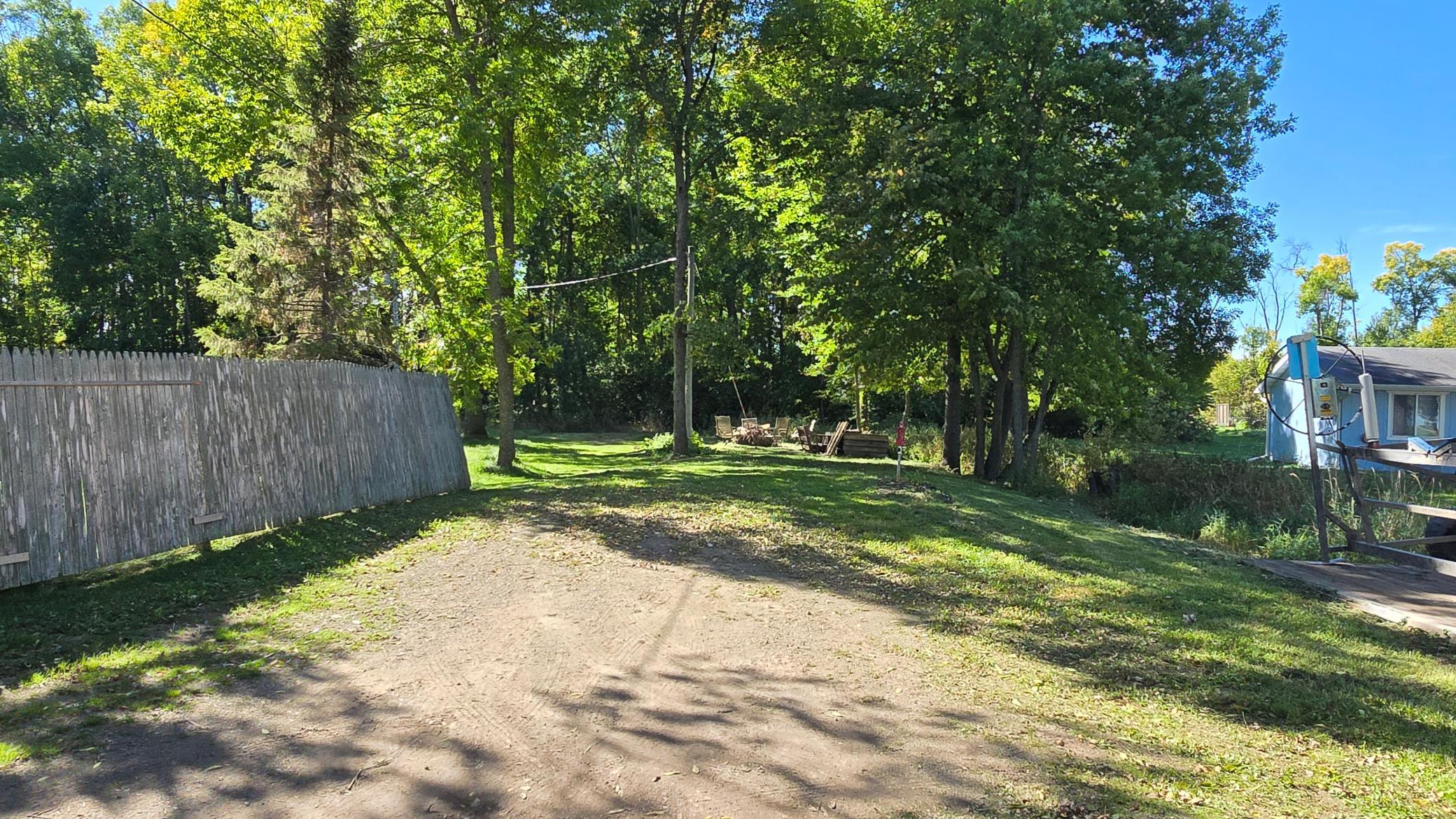 Tree-shaded yard with a weathered privacy fence, grassy lawn, dirt drive, and outdoor seating.