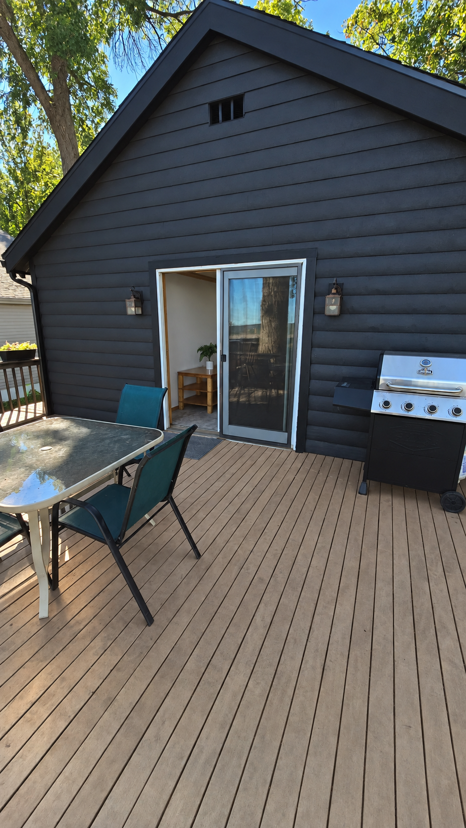 Wooden deck with outdoor dining set and gas grill beside a dark-sided home.