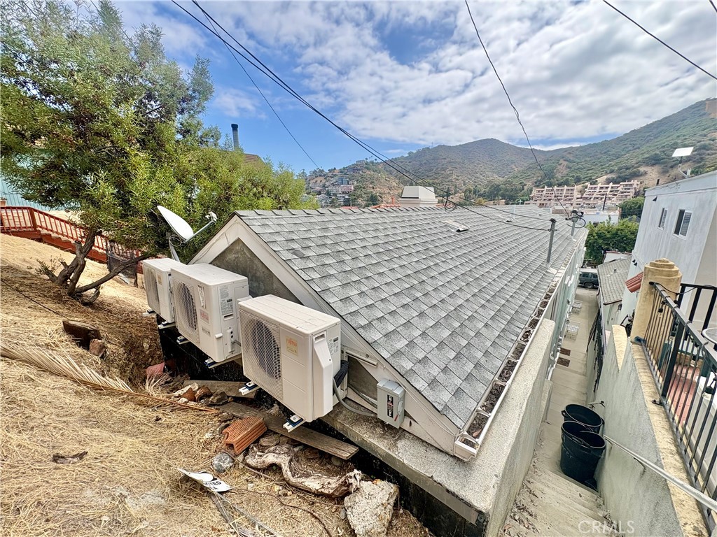 Sloped shingle roof with outdoor air-conditioning units beside a hillside walkway.