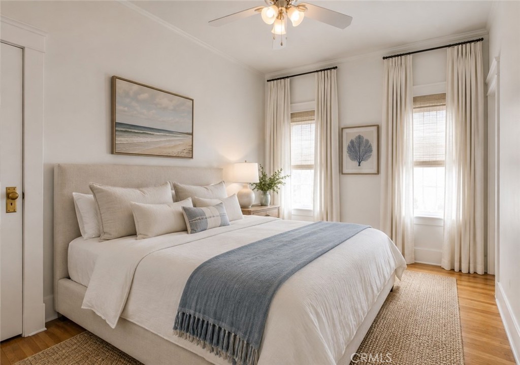 Bright bedroom with a white bed, blue throw, tall curtained windows, and coastal artwork.