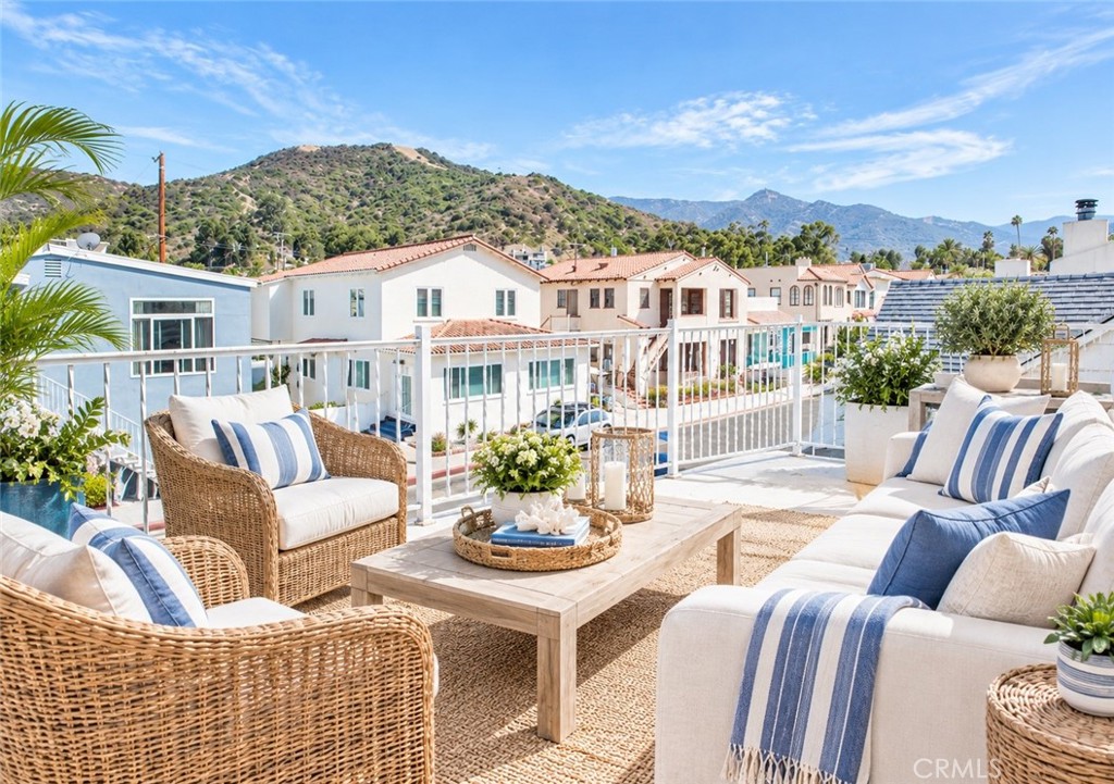 Rooftop terrace with wicker seating and blue-striped cushions overlooking homes and green hills.