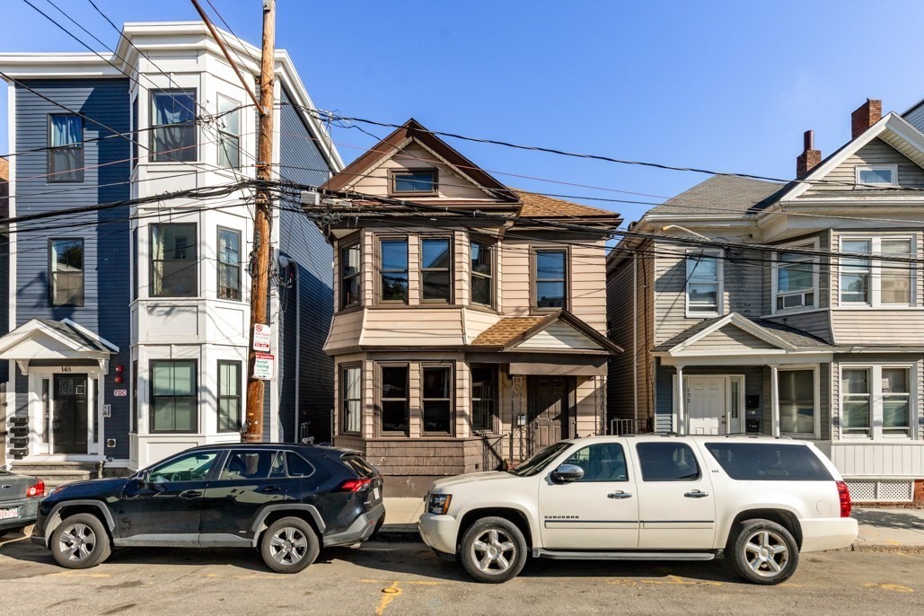 Tan multi-story Boston home with a front porch, between neighboring houses on a sunny street.