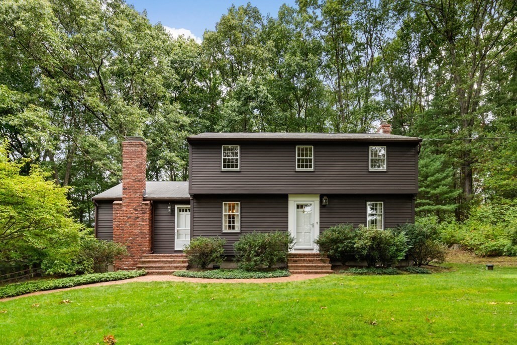 Two-story brown home with a brick chimney, white entry doors, and a tree-lined lawn.