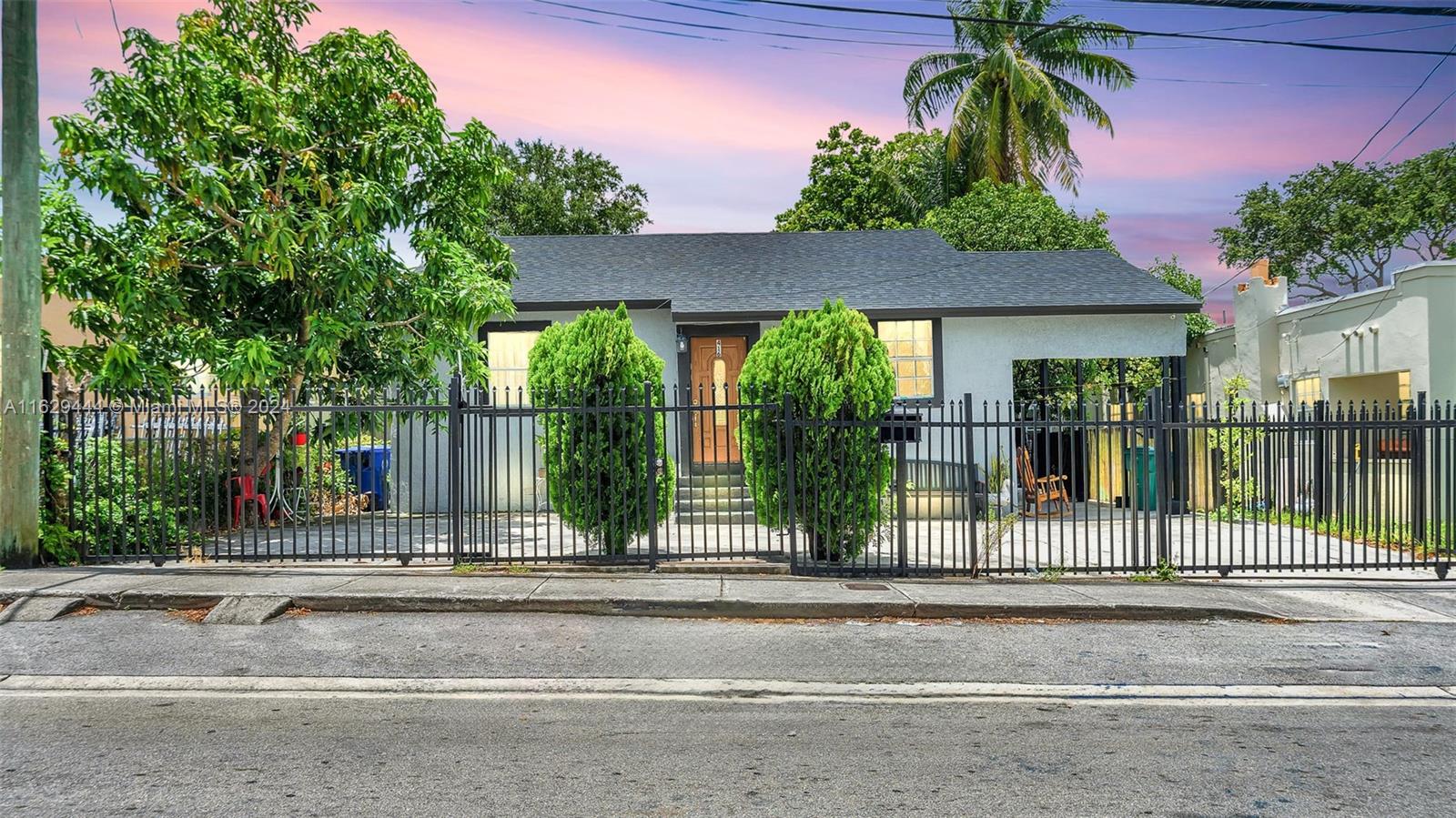 Single-story home behind a black metal fence, framed by tropical landscaping beneath a pink dusk sky.