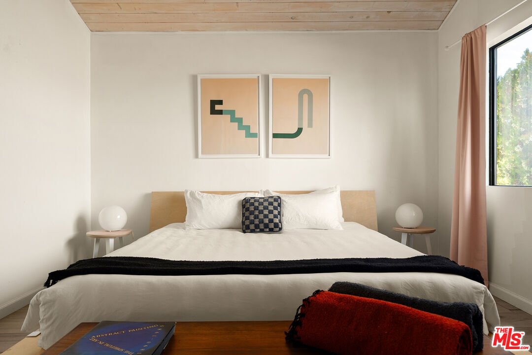 Staged bedroom with a white bed, plank ceiling, abstract prints, and a window with a peach curtain.