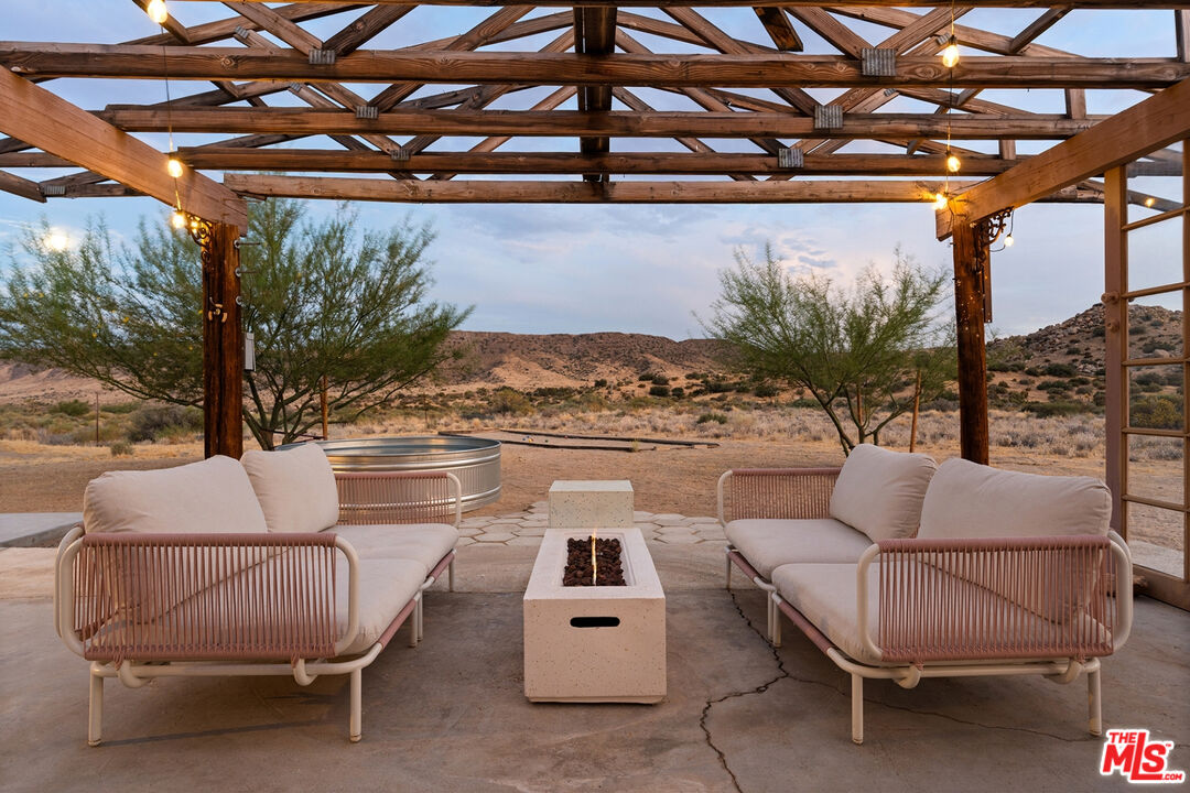 Pergola-covered patio with cushioned seating, a fire table, and hot tub overlooking desert hills.