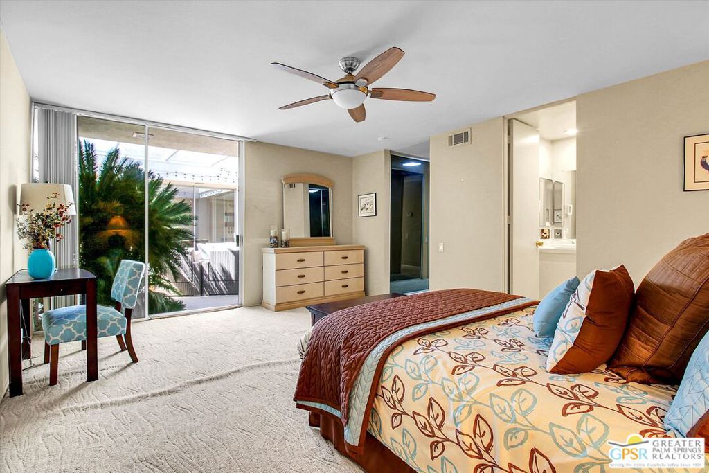 Furnished bedroom with a patterned bedspread, ceiling fan, and sliding glass doors overlooking palm foliage.