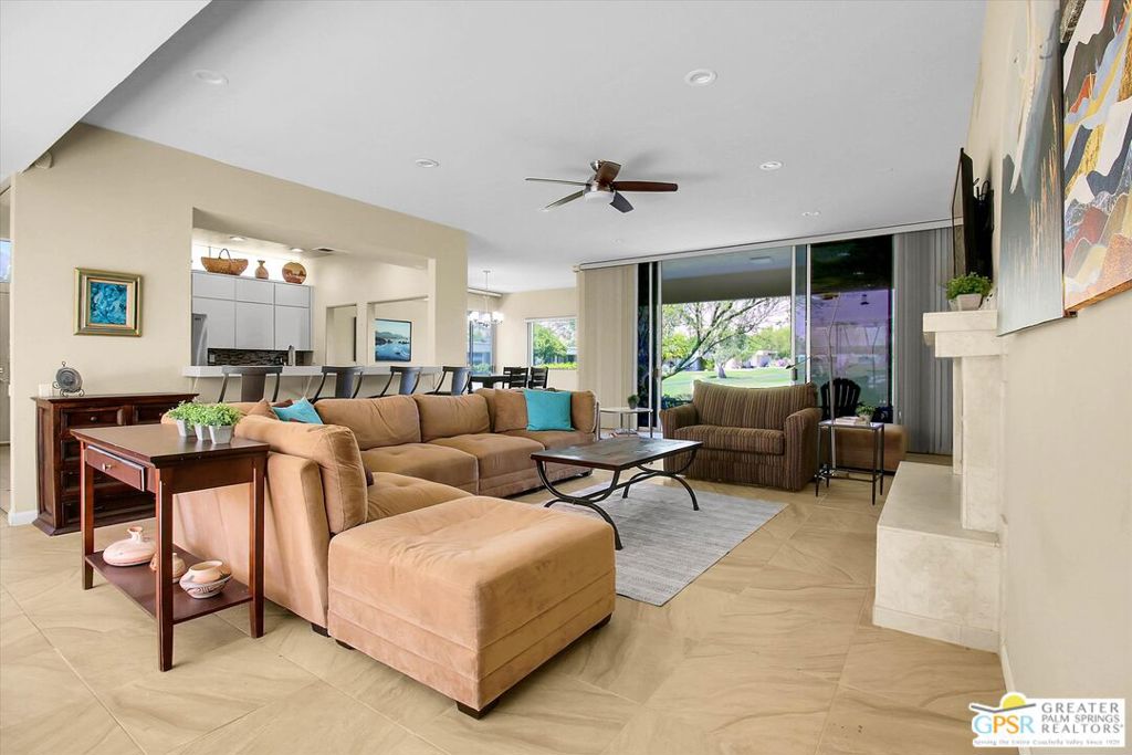 Furnished living room with tan sectional, tiled floors, fireplace, and sliding glass doors overlooking a lawn.