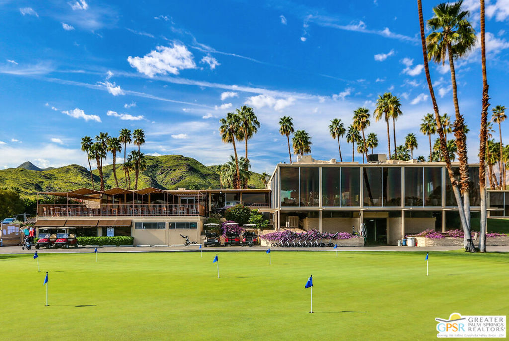 Golf course putting green and modern clubhouse framed by palm trees, with mountains behind.
