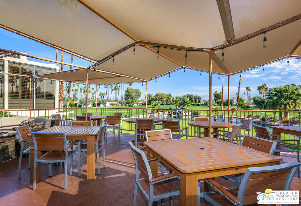 Covered outdoor dining patio with wooden tables and chairs overlooking green grounds and palm trees.