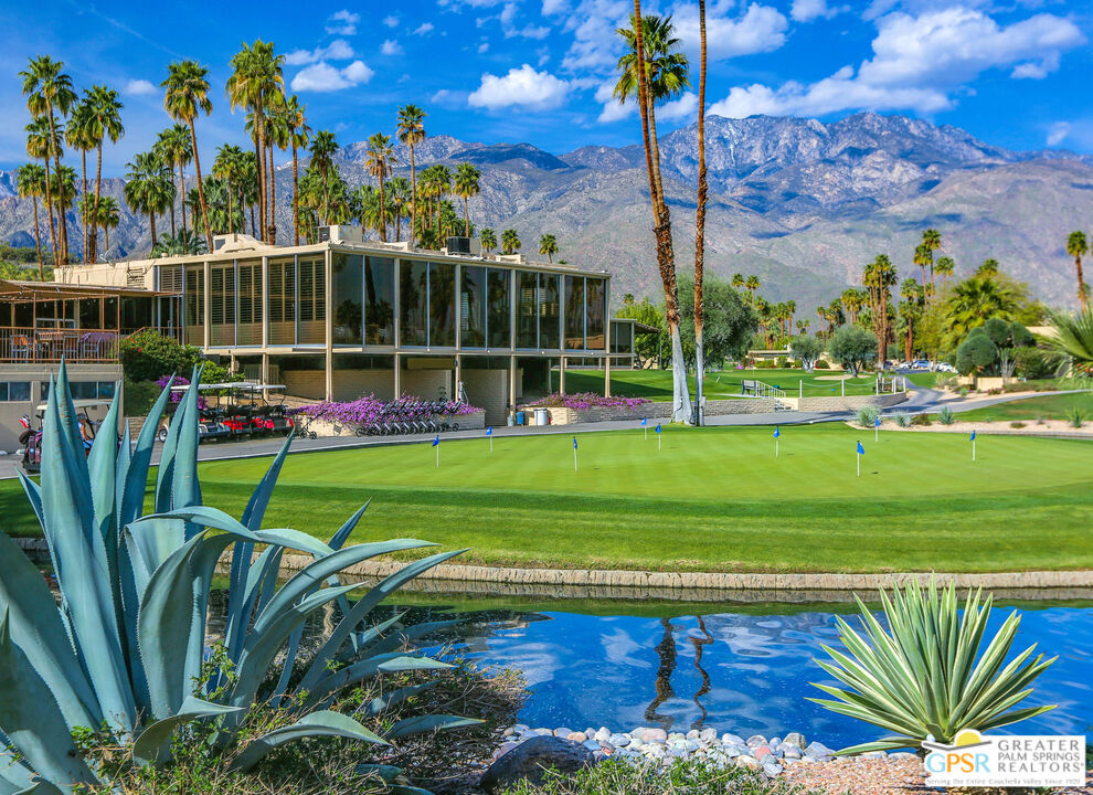 Modern glass-walled home beside a golf green and pool, with palm trees and mountains behind it.