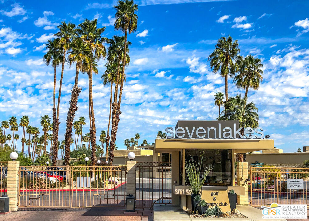 Seven Lakes community entrance with a gated drive, guard booth, and tall palm trees.
