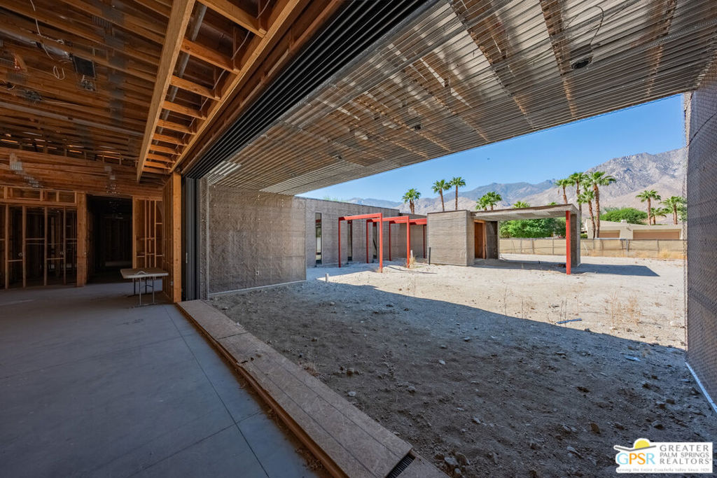 Unfinished home interior opens onto a dirt courtyard with red steel posts, palm trees, and mountains.