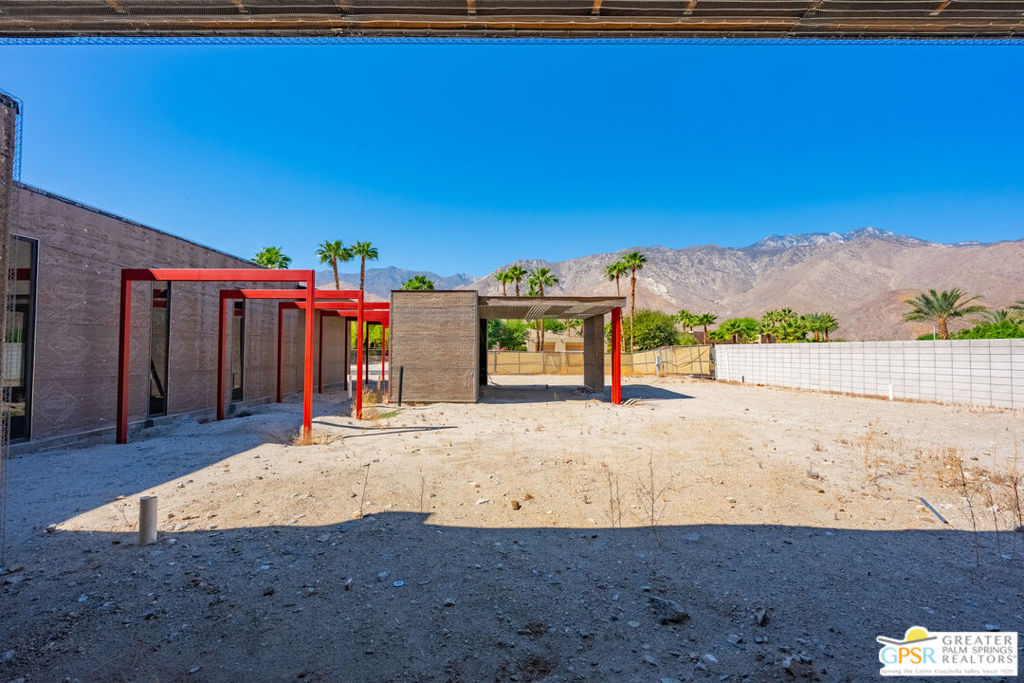 Modern home under construction with red steel frames, a bare yard, palm trees, and mountains beyond.
