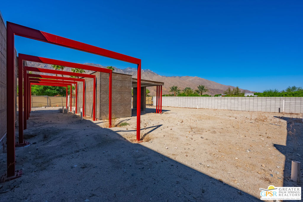 Unfinished property with red steel framing and a sandy lot, backed by mountains.
