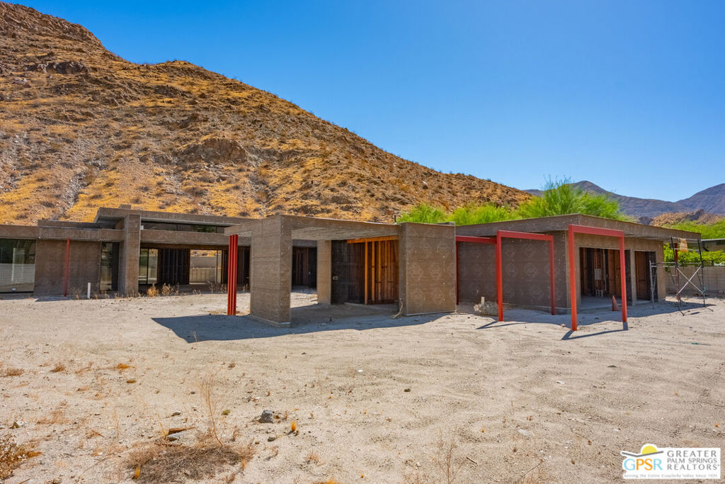 Single-story home under construction, with exposed concrete walls and red steel framing against a desert mountain.