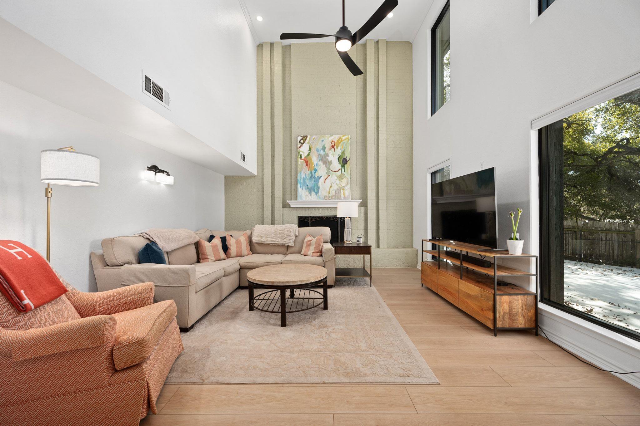Vaulted living room with a pale brick fireplace wall, beige sectional, and large windows overlooking a fenced yard.
