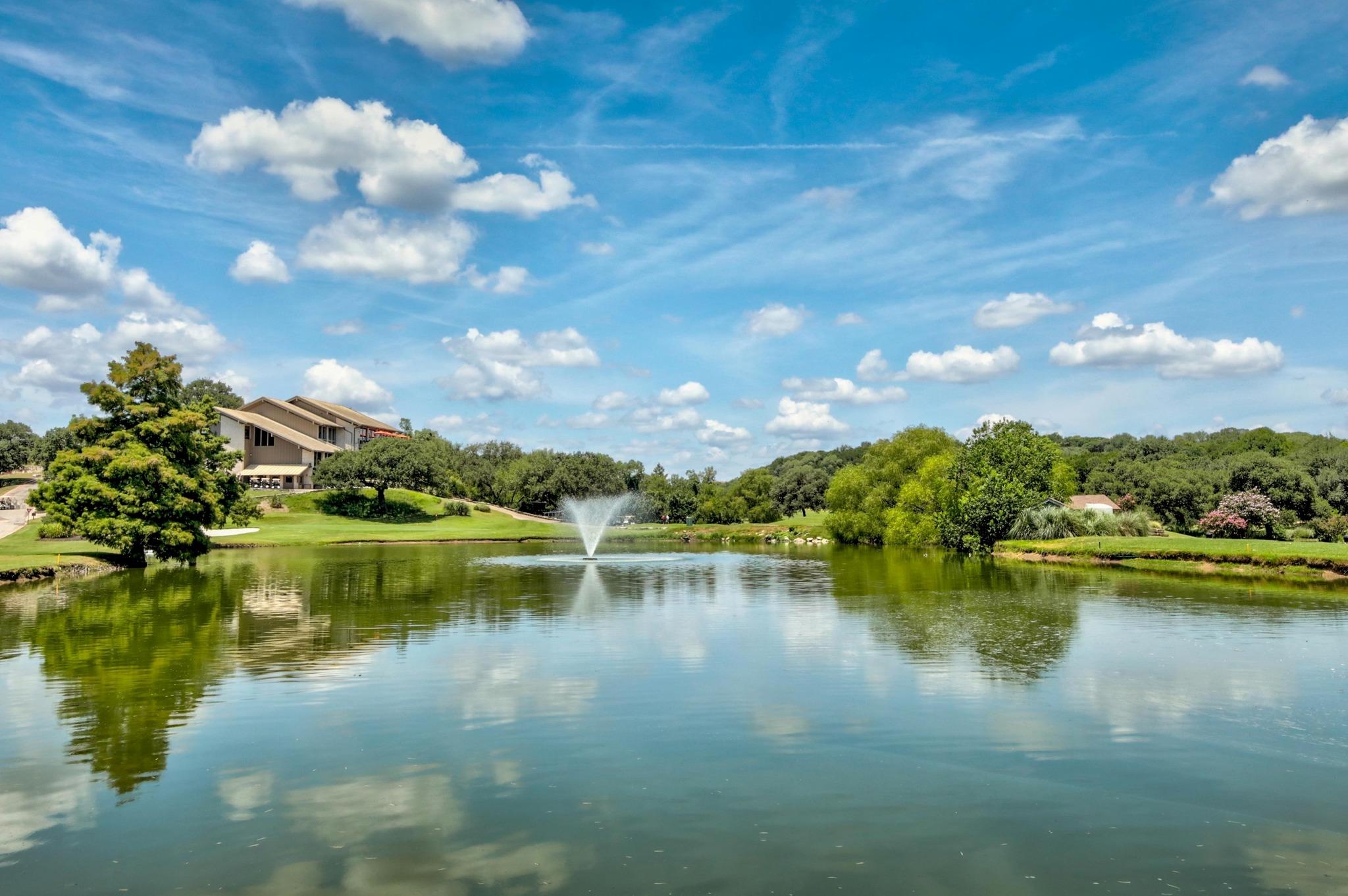 Landscaped pond with a fountain, tree-lined banks, and a large residence in the background.