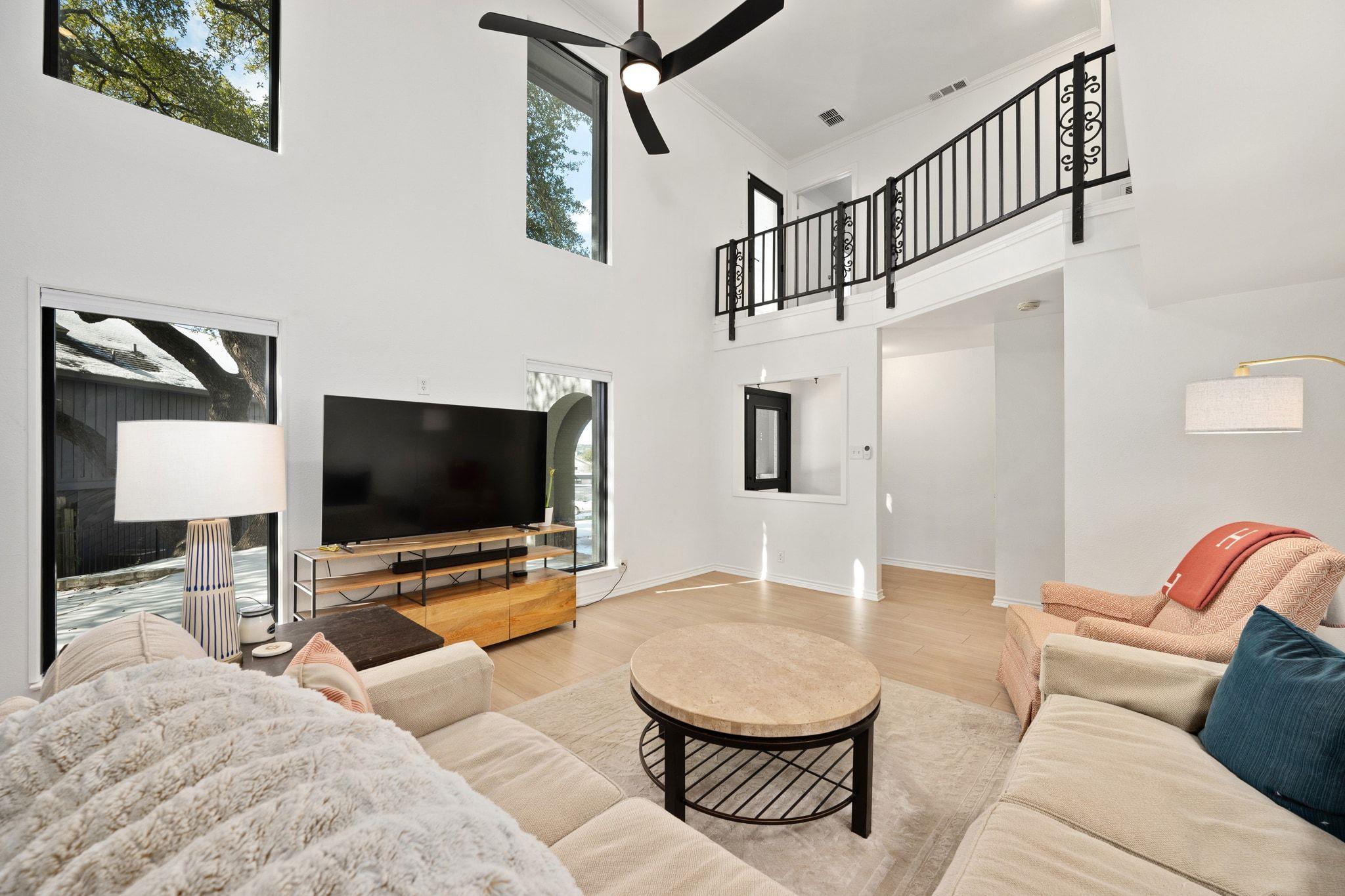 Double-height living room with large windows, loft railing, and neutral seating around a round coffee table.