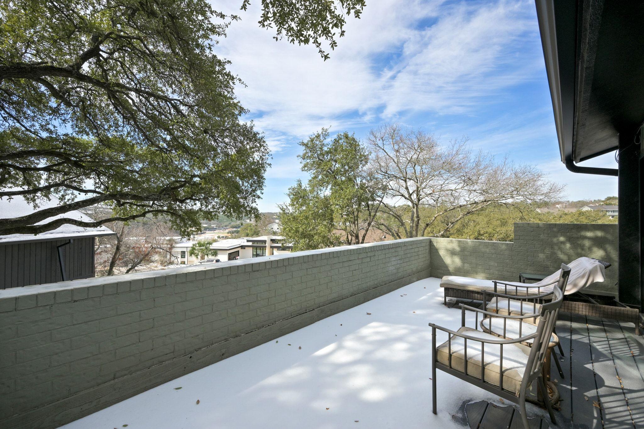 Snow-covered deck with cushioned seating, a brick wall, and views of trees and nearby homes.