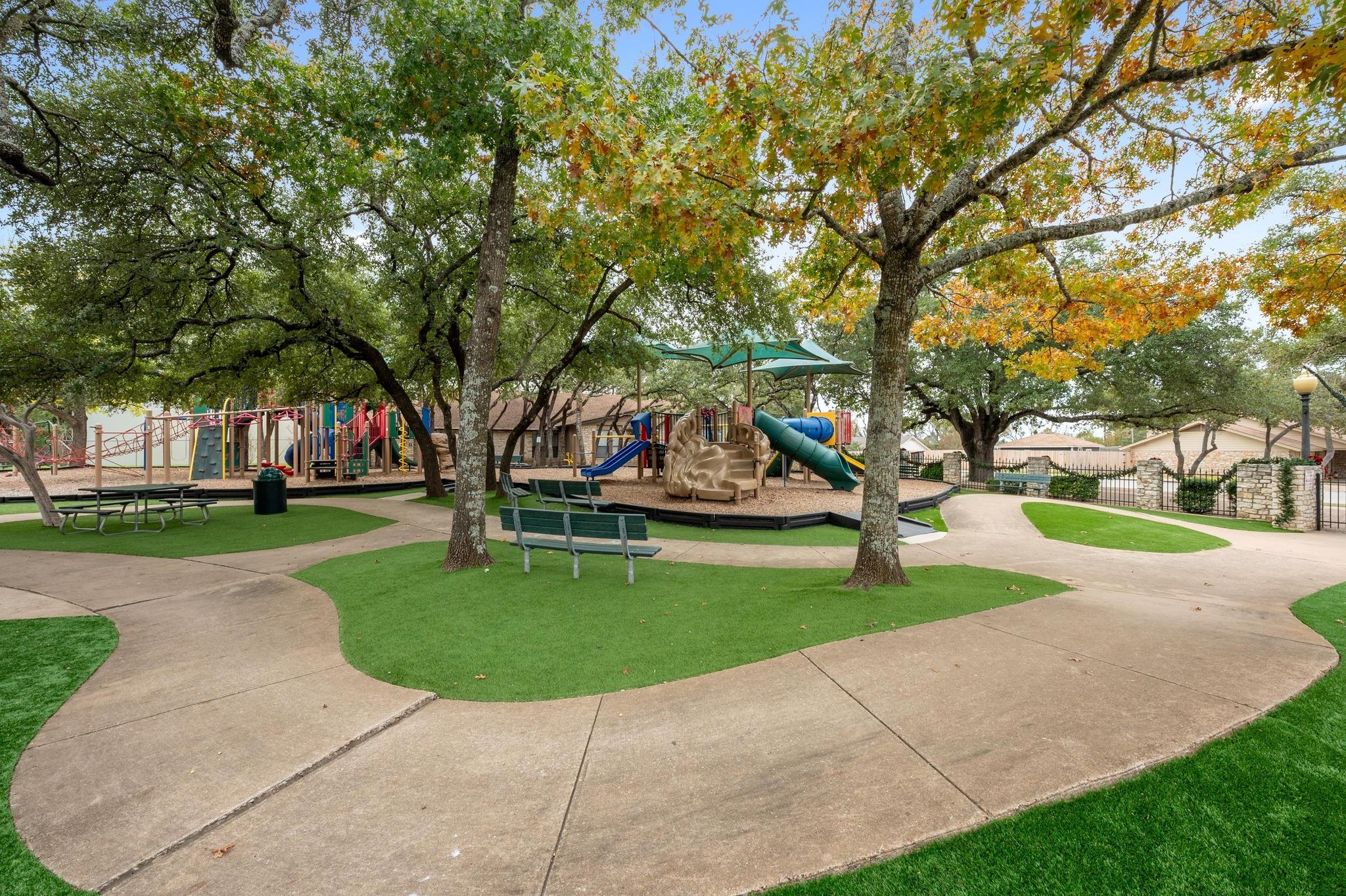 Community playground with colorful slides and climbing equipment beneath mature trees, beside benches and paved paths.