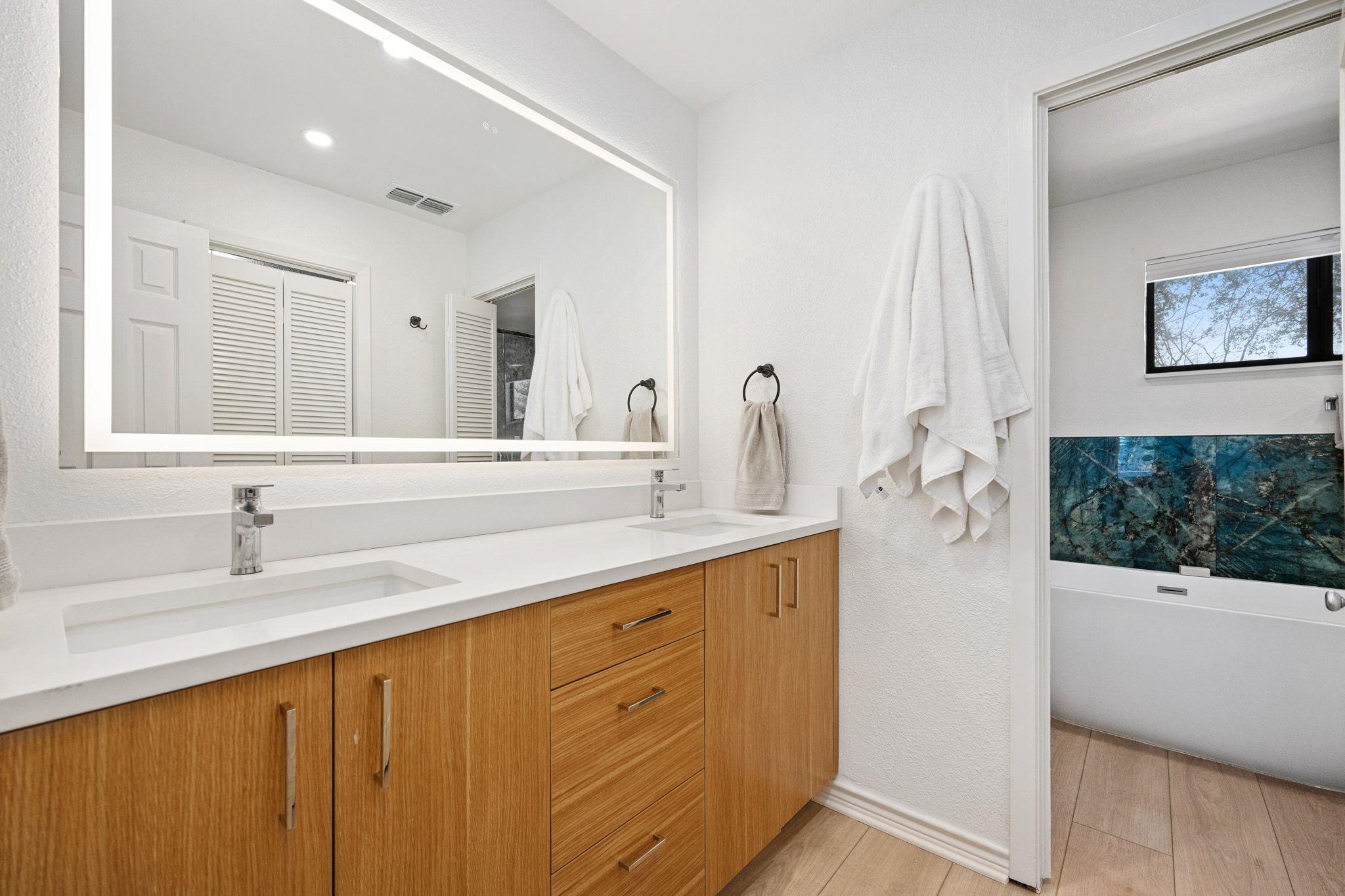 Bathroom vanity with double sinks, wood cabinets, illuminated mirror, and a tub with blue-green tile.