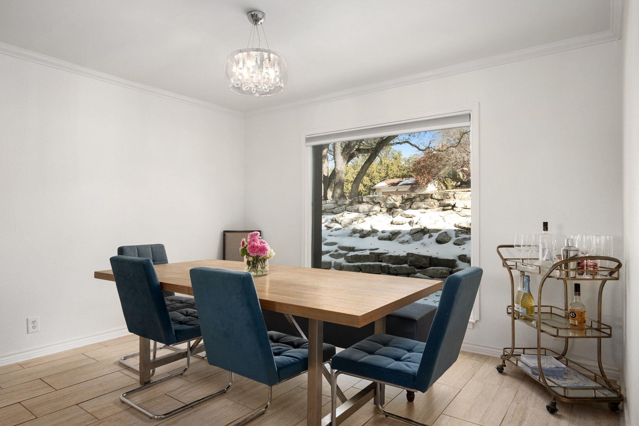 Dining room with a wooden table, navy chairs, chandelier, and window overlooking a stone wall.