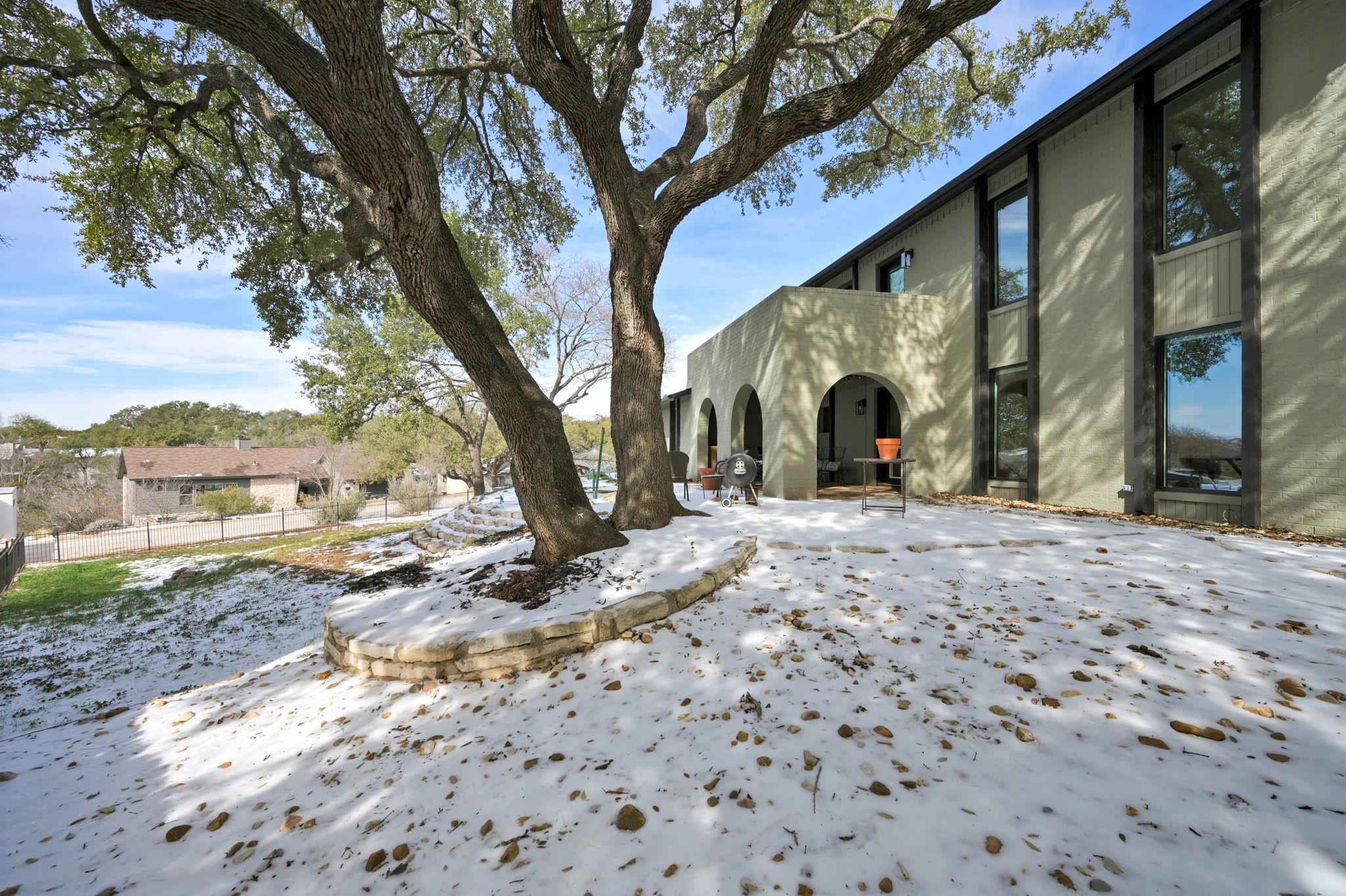 Snow-covered patio beside a pale brick home, shaded by a large tree and lined with arched openings.