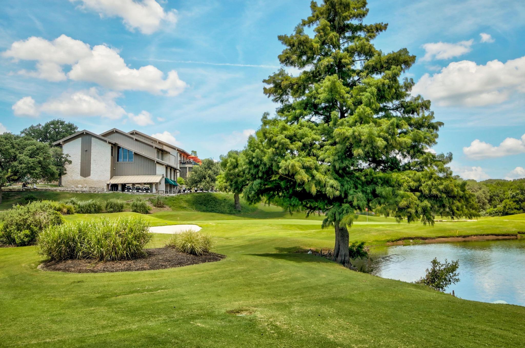 Golf course with a pond and large tree beside a multi-story building.