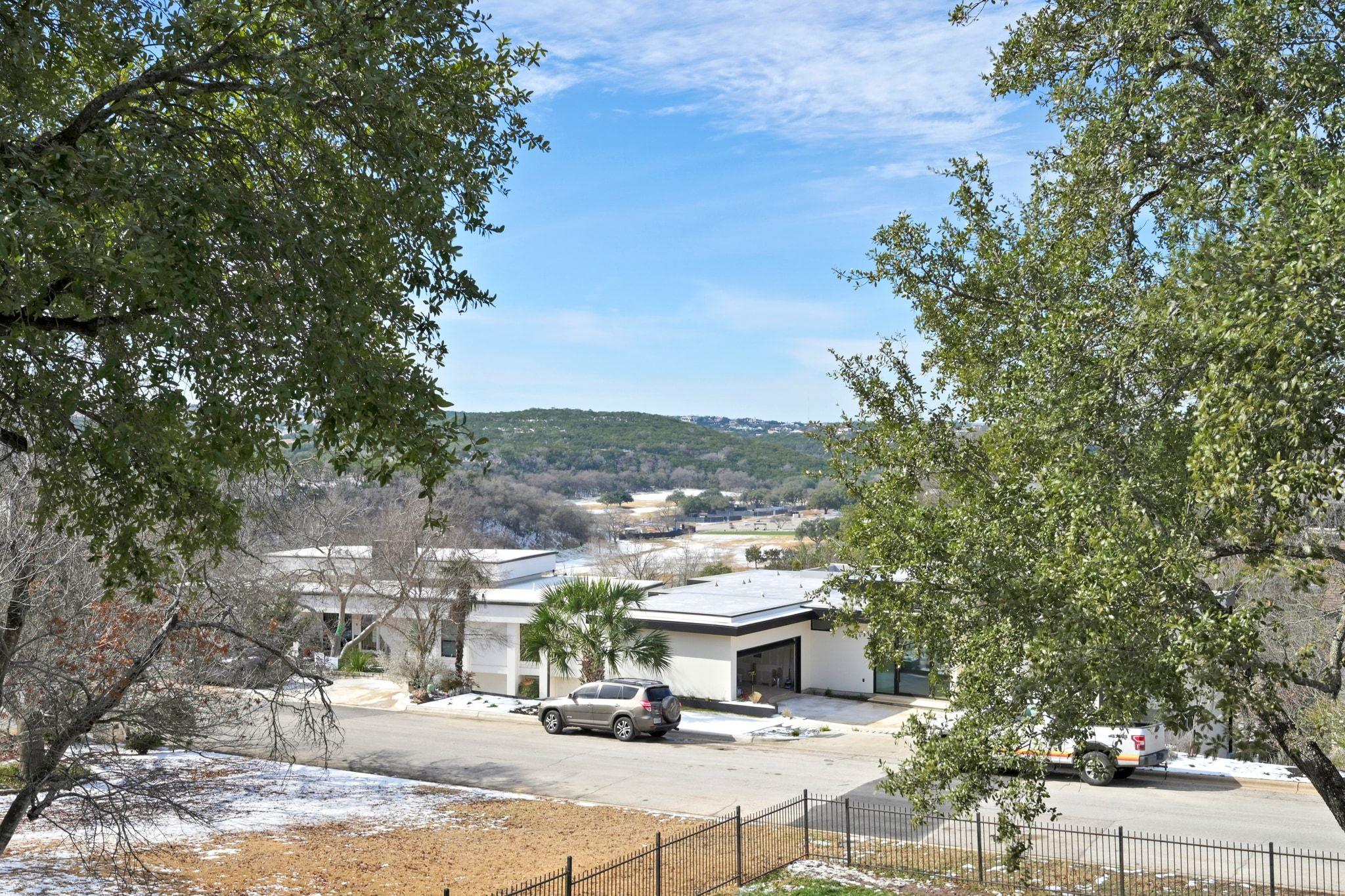 Modern white home across a street, framed by trees with wooded hills beyond.